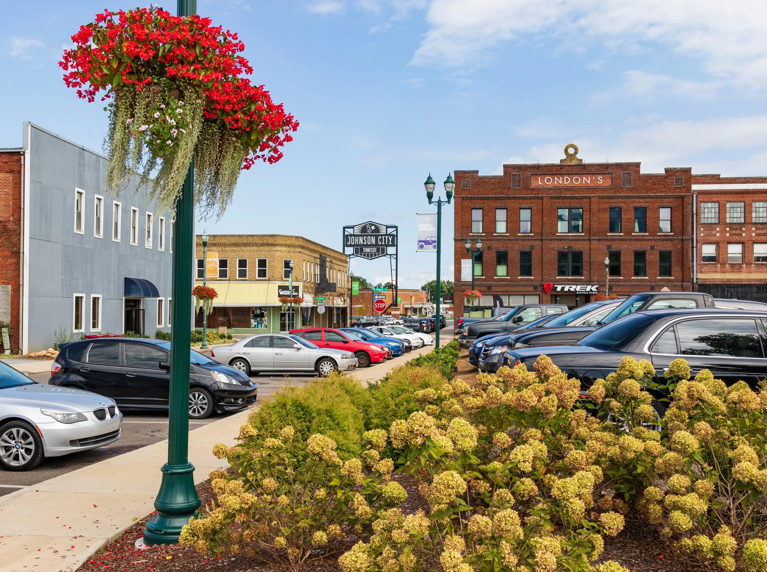 A downtown street scene with parked cars, brick and storefront buildings, hanging flower baskets, and a prominent sign for Johnson City, set against a partly cloudy sky.