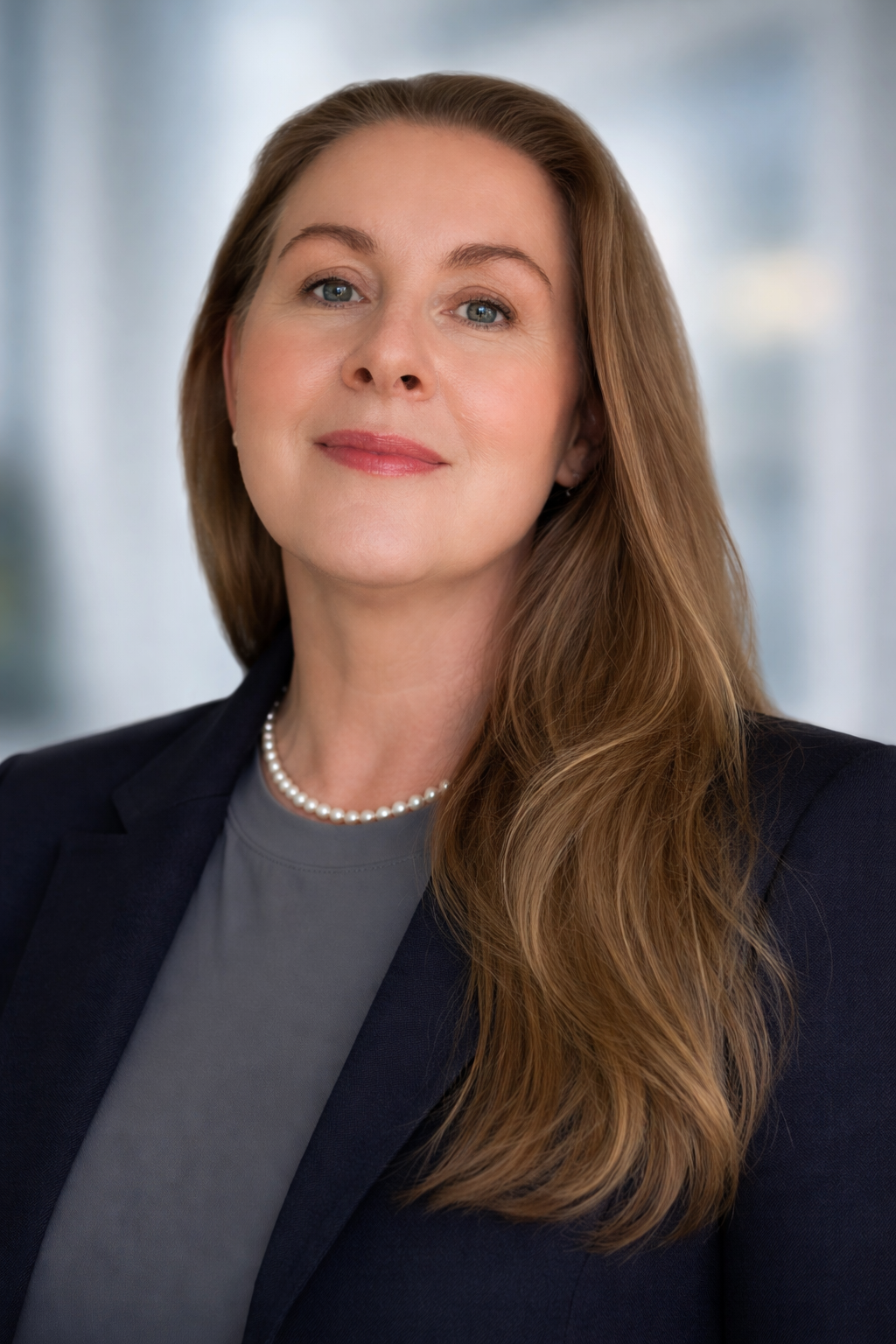 Professional woman with long, wavy red hair and blue eyes, wearing a dark blazer, gray top, pearl necklace, and light makeup, smiling softly in a blurred office background.