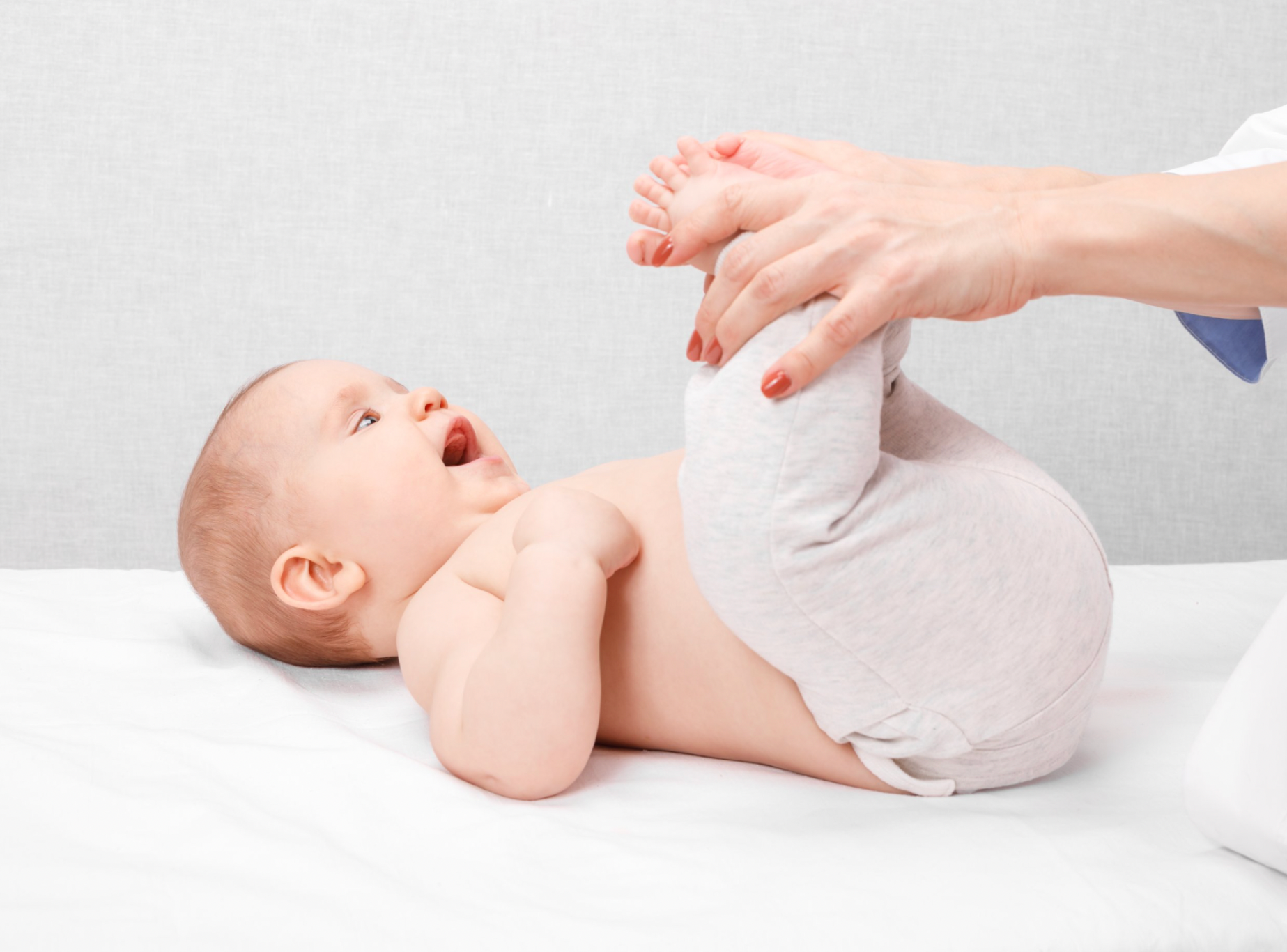 Bebê deitado na cama, brincando com os pés, enquanto uma pessoa segura suas pernas.