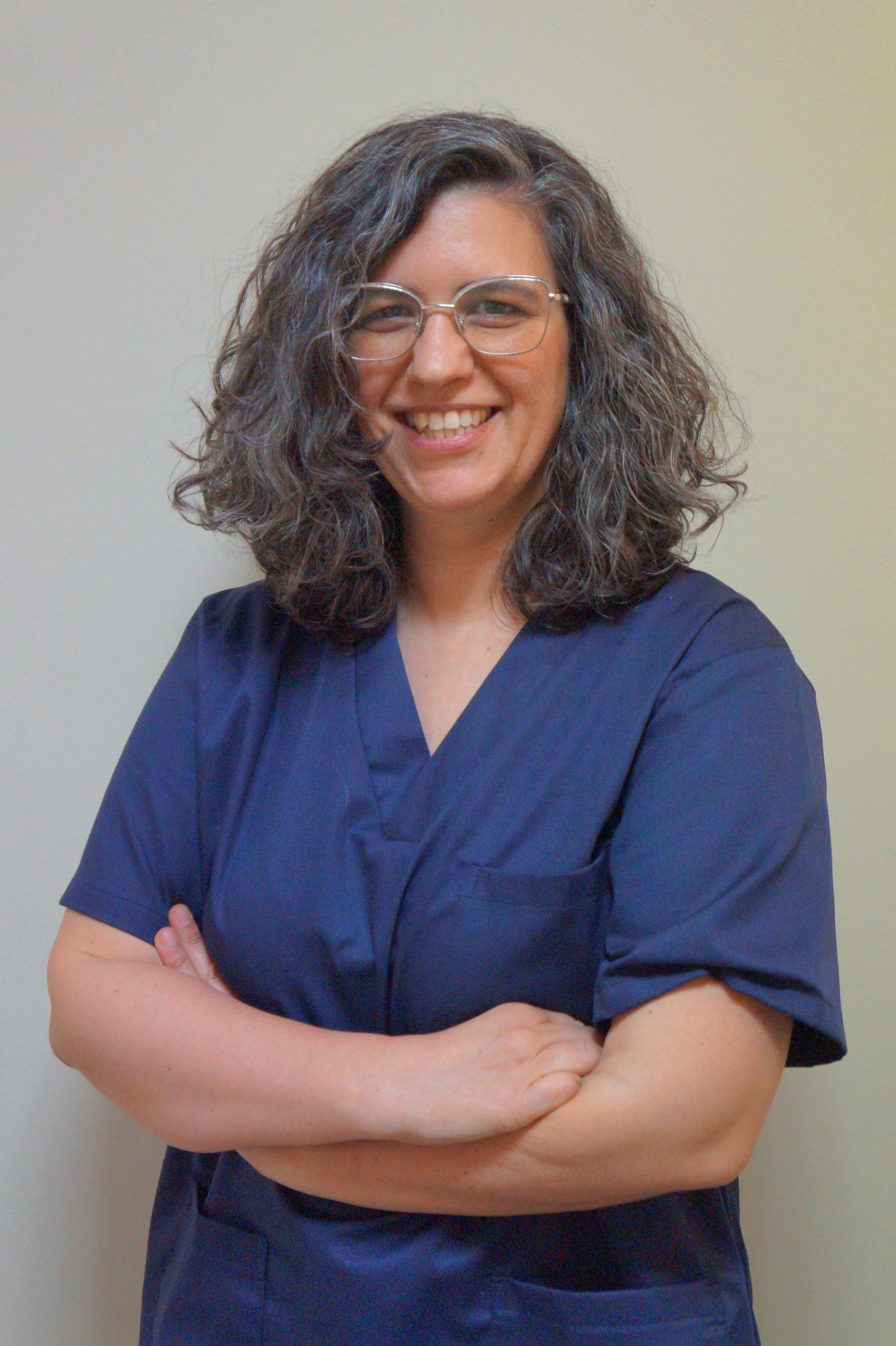 Médica da equipa sorridente com cabelos cabelos aos caracóis e óculos, com uniforme azul vestido , de braços cruzados, a pousar contra fundo de parede clara.