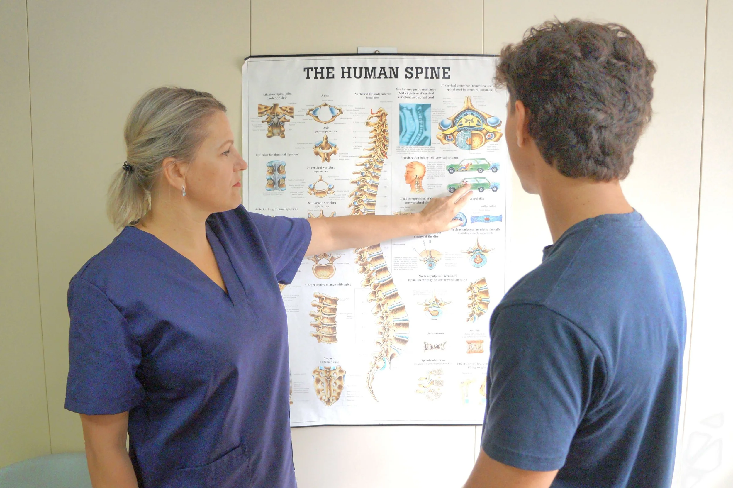 Profissional de saúde explicando o funcionamento da coluna vertebral a um jovem na frente de um pôster anatômico que mostra a coluna humana.