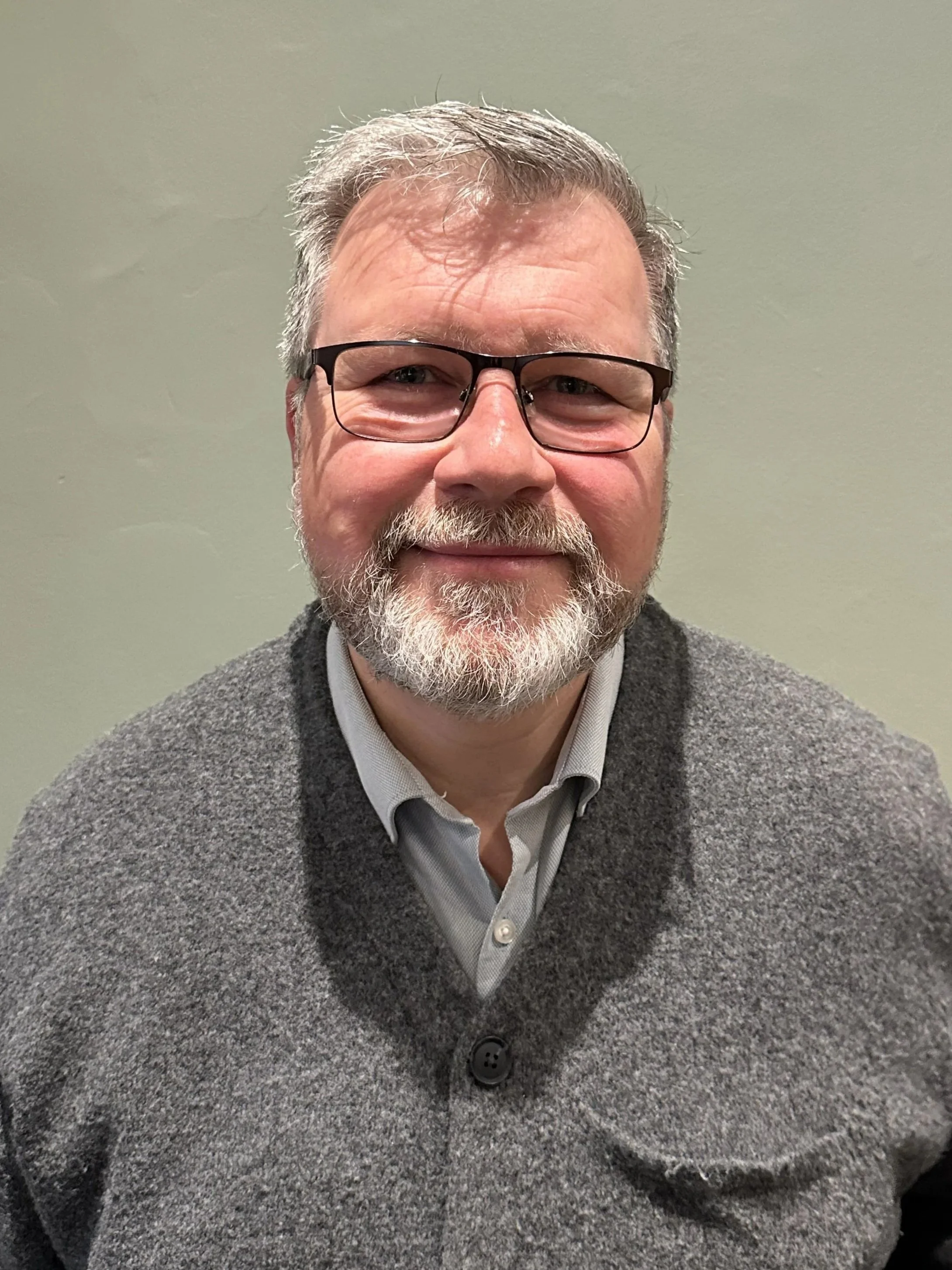 Image of man with a beard and glasses, with a grey jumper standing in front of a green wall.