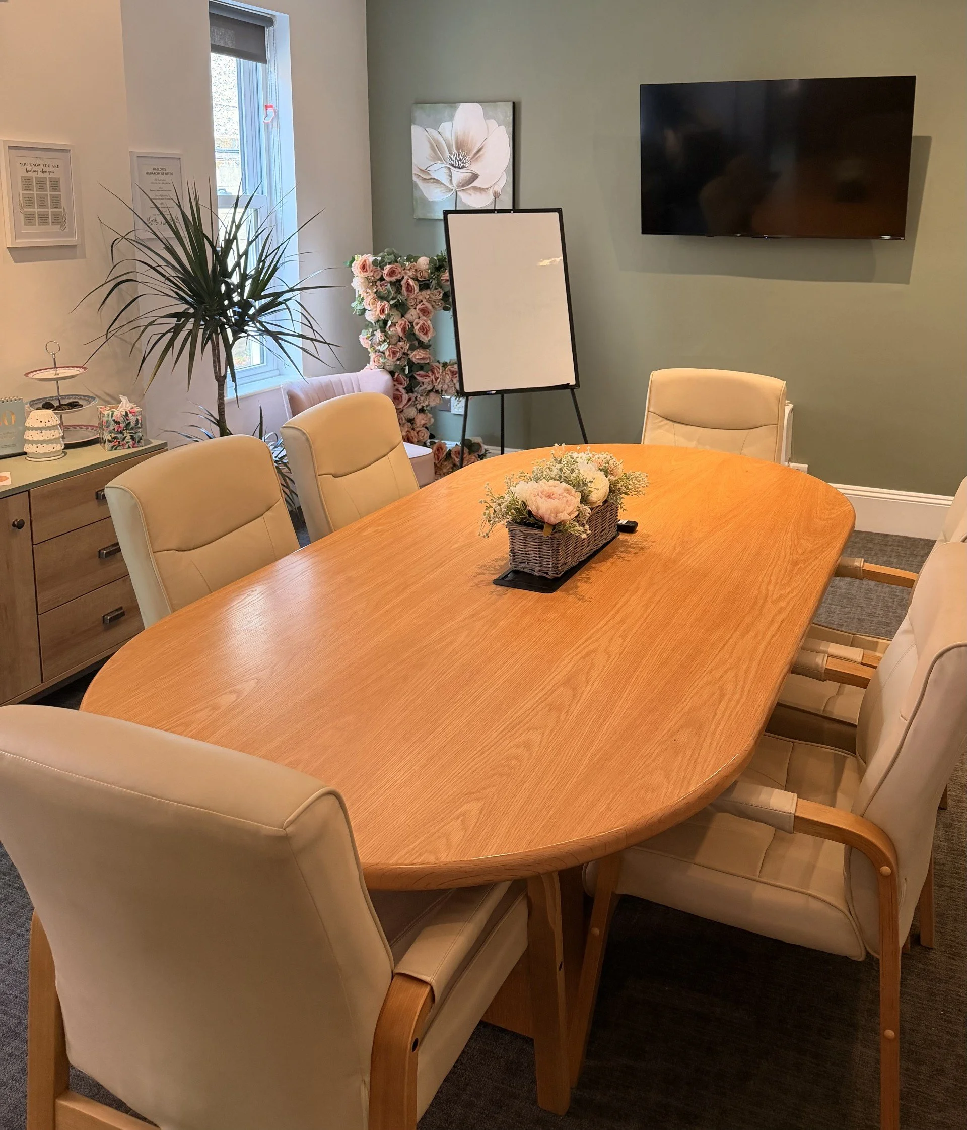 A wooden conference style table with six cream chairs around it and a floral arrangement in the centre of the table. TV on the wall of the meeting space.