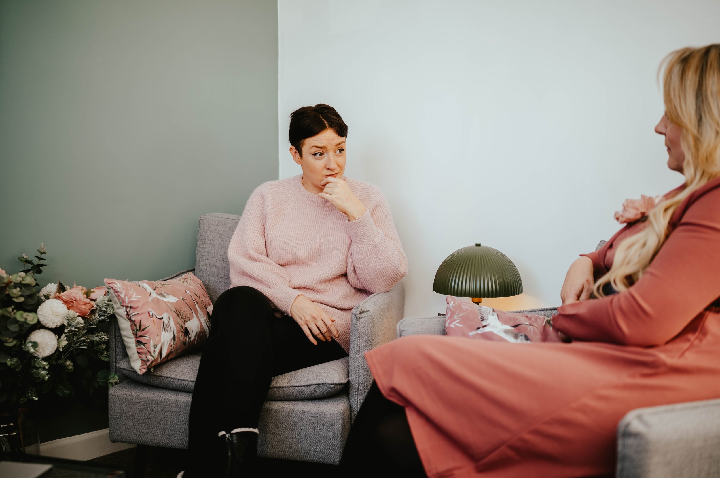 Lady with short, dark hair in a pink jumper talking to a lady with long, blonde hair and a bright pink dress in a therapy environment.