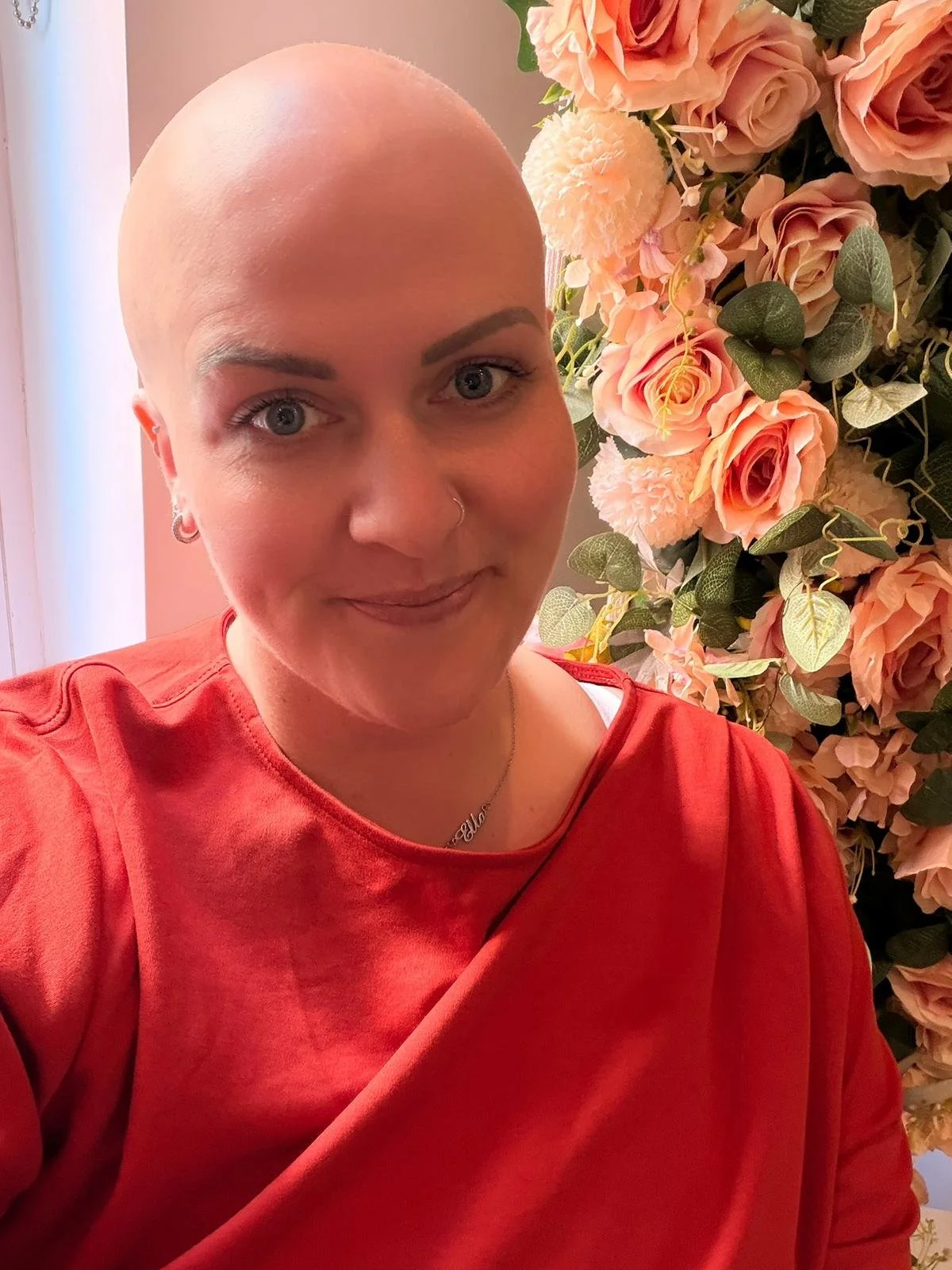A smiling woman with no hair, wearing an orange top with flowers in the background.