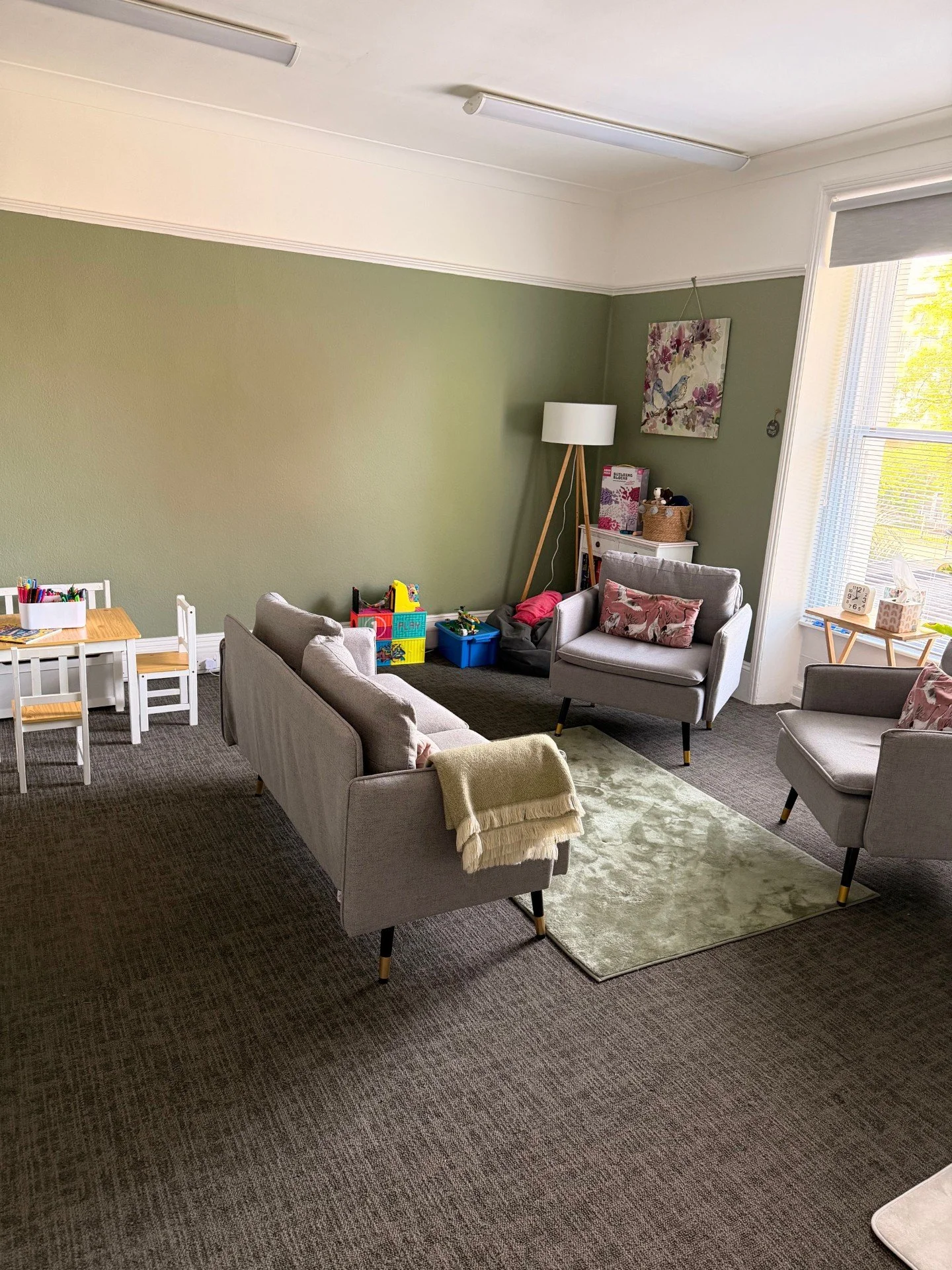 Therapy room with green walls, a grey sofa and two chairs. Toys and art supplies around the room for children to use during sessions.