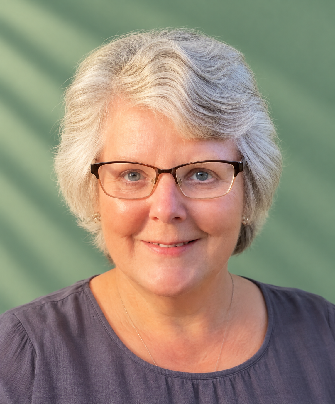 A smiling older woman with short gray hair and glasses, outdoors with green foliage and yellow flowers in the background.