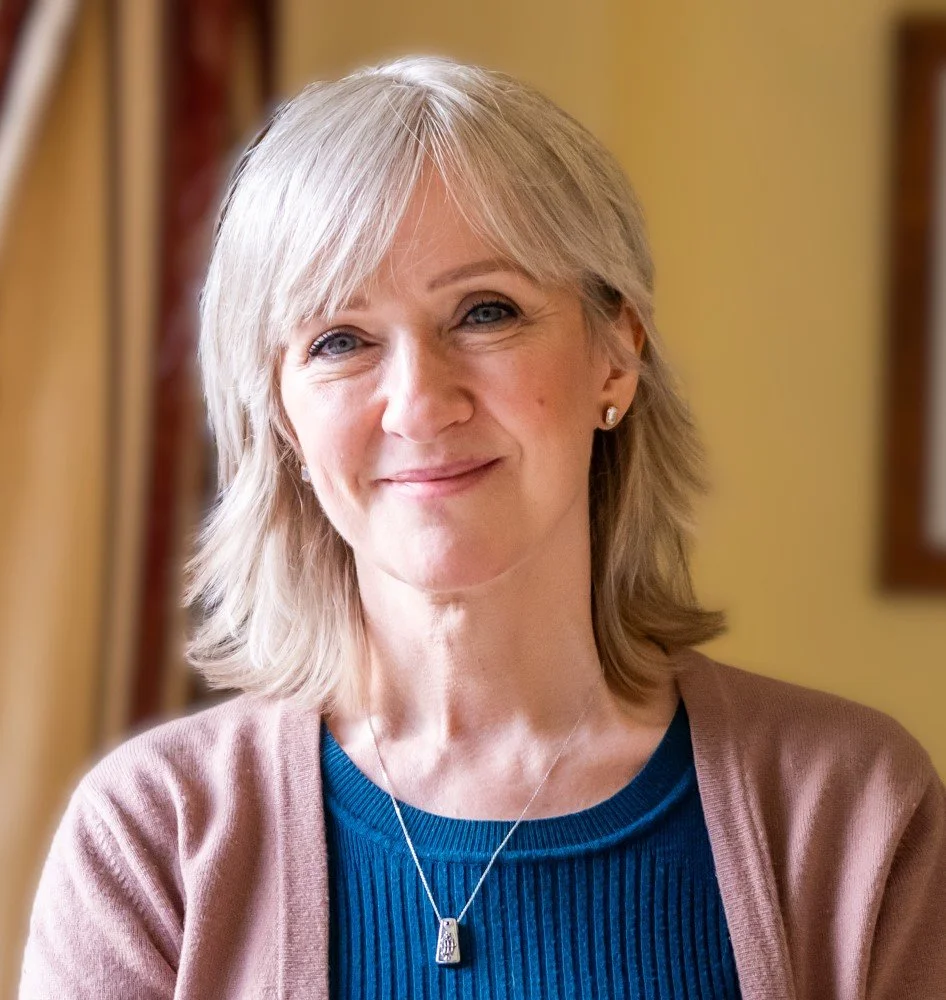 Smiling lady in a blue top and pink cardigan Blonde shoulder length hair.