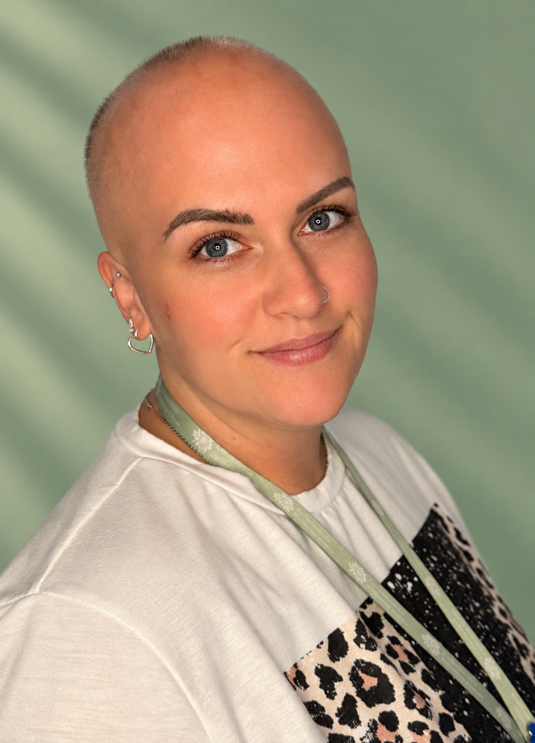 A smiling woman with no hair, wearing an orange top with flowers in the background.