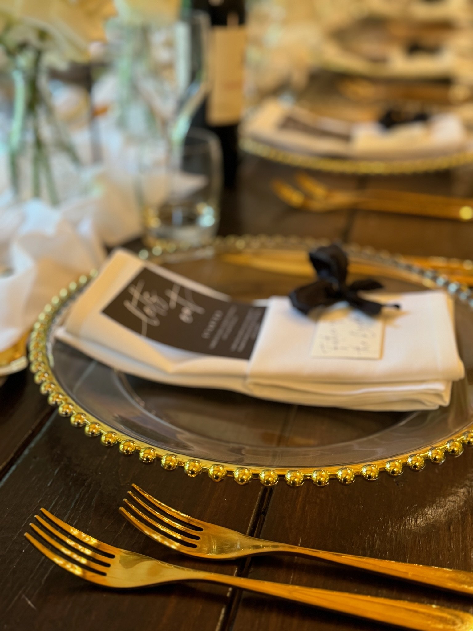 Elegant table setting with gold-trimmed glass charger plate, white napkin, black ribbon, black menu, gold cutlery, and wine glass, on a dark wooden table
