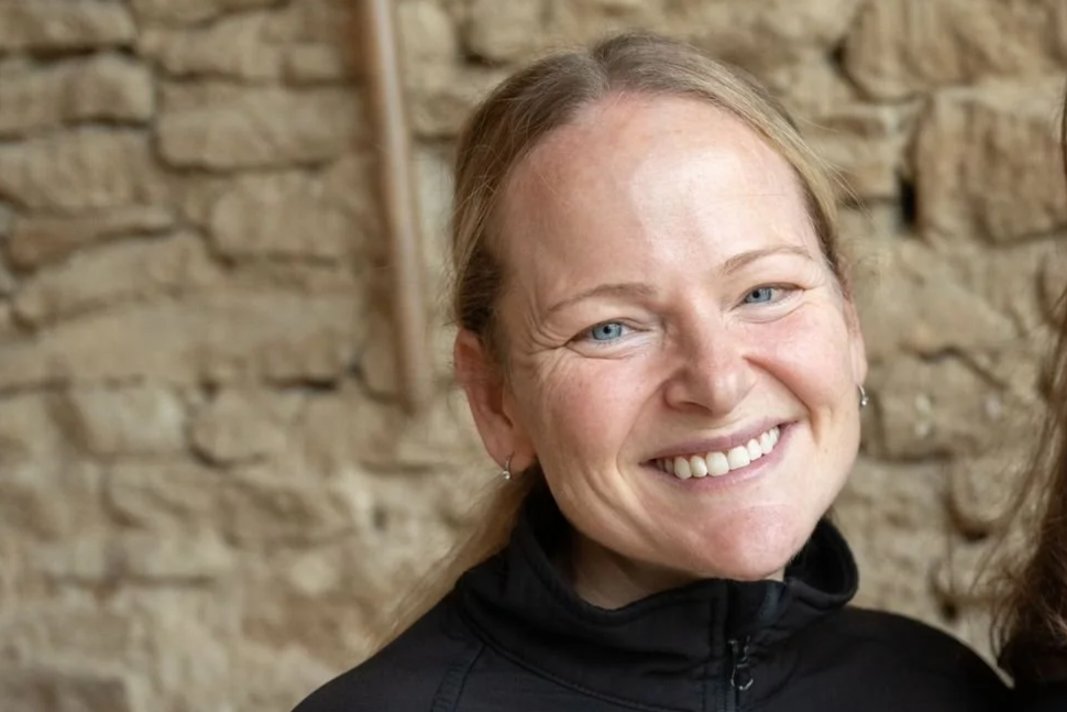 A smiling woman with curly hair, wearing glasses and hoop earrings, in an indoor setting.