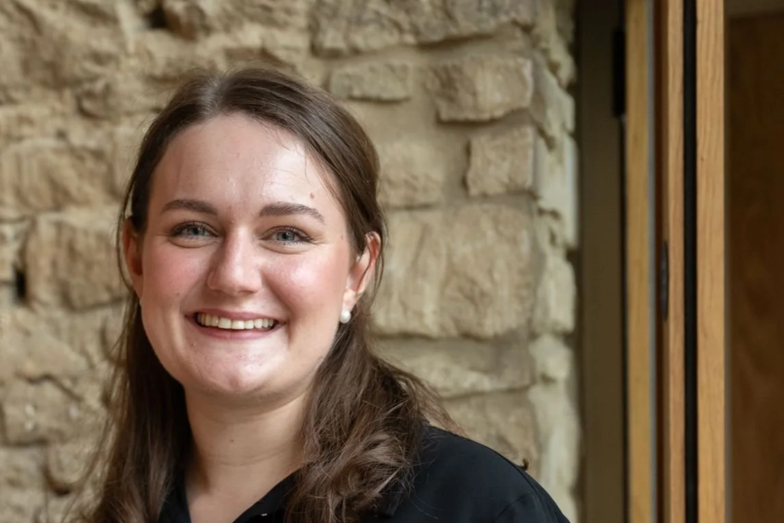 A young woman with shoulder-length black hair smiling in a greenhouse with green plants around her.
