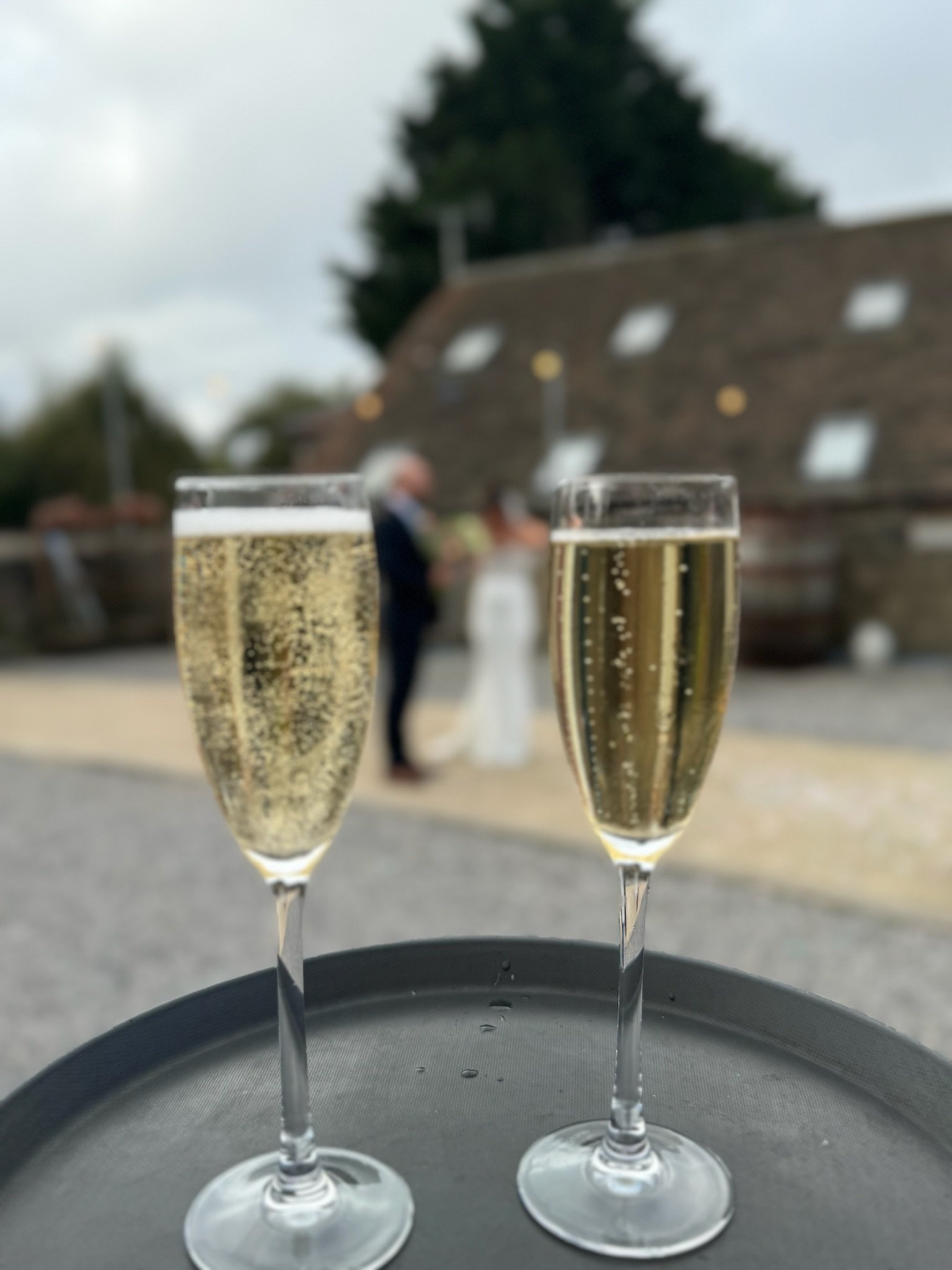 Two glasses of champagne on a black tray with a blurred outdoor wedding scene in the background.