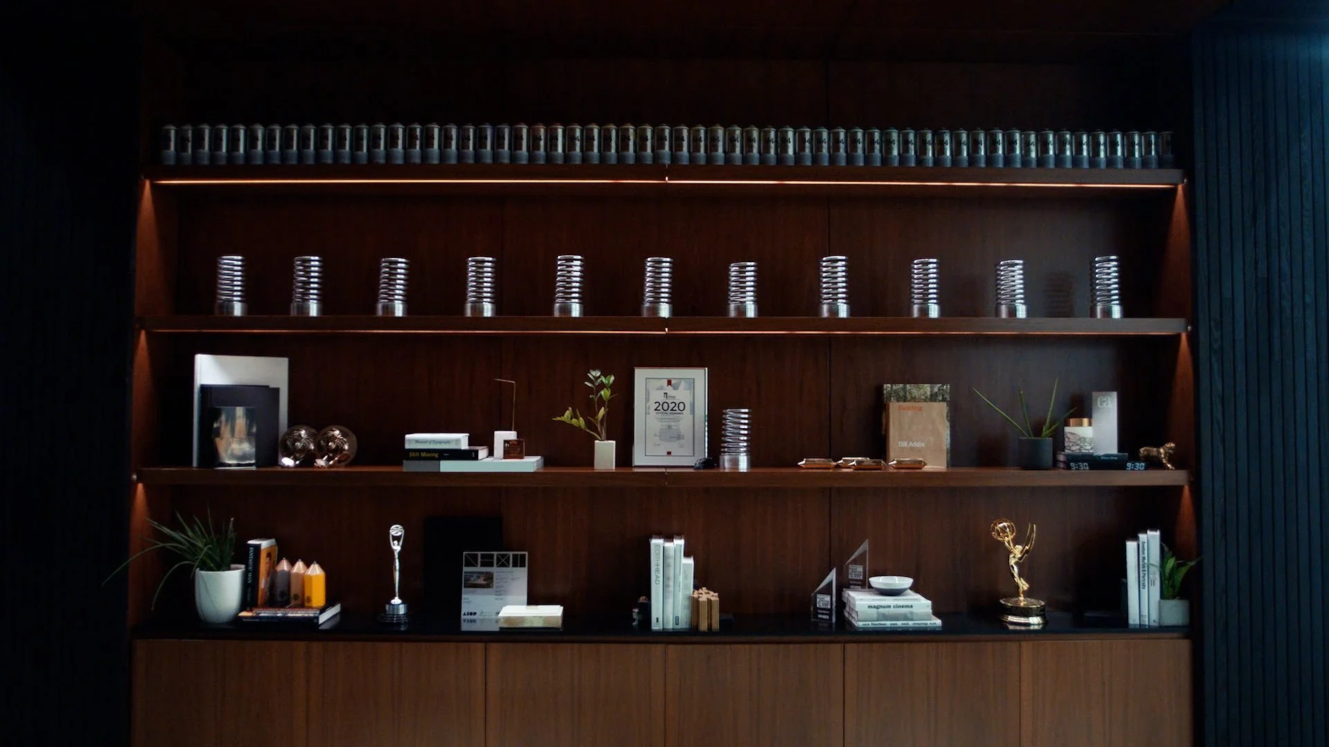 Wooden bookshelves with decorative items, awards, books, potted plants, and framed certificates or awards inside a modern office or lobby.