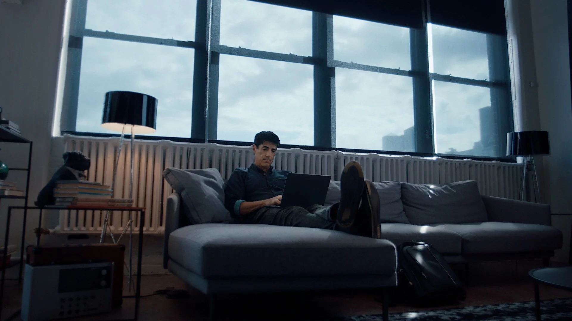 Man sitting on a gray sectional sofa using a laptop in a living room with large windows showing a cloudy sky