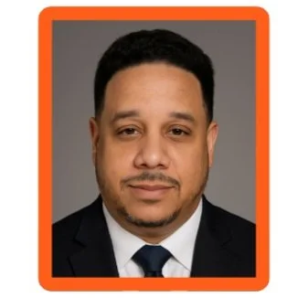 Professional headshot of a man with short black hair, wearing a dark suit, white shirt, and dark tie, against a gray background with an orange border.