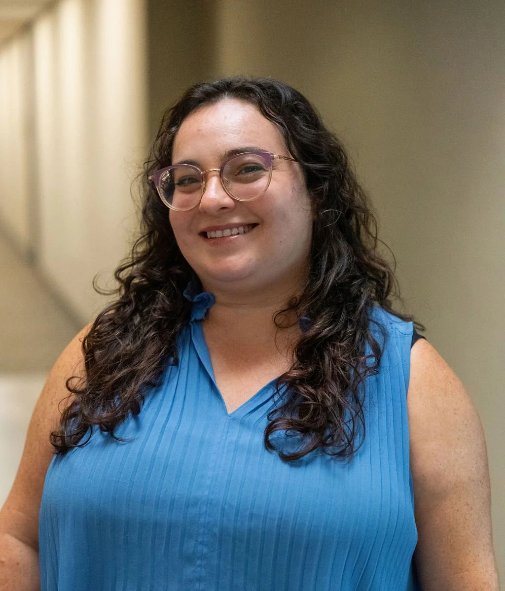 A woman with long curly dark hair, wearing glasses and a sleeveless blue top, smiling indoors.