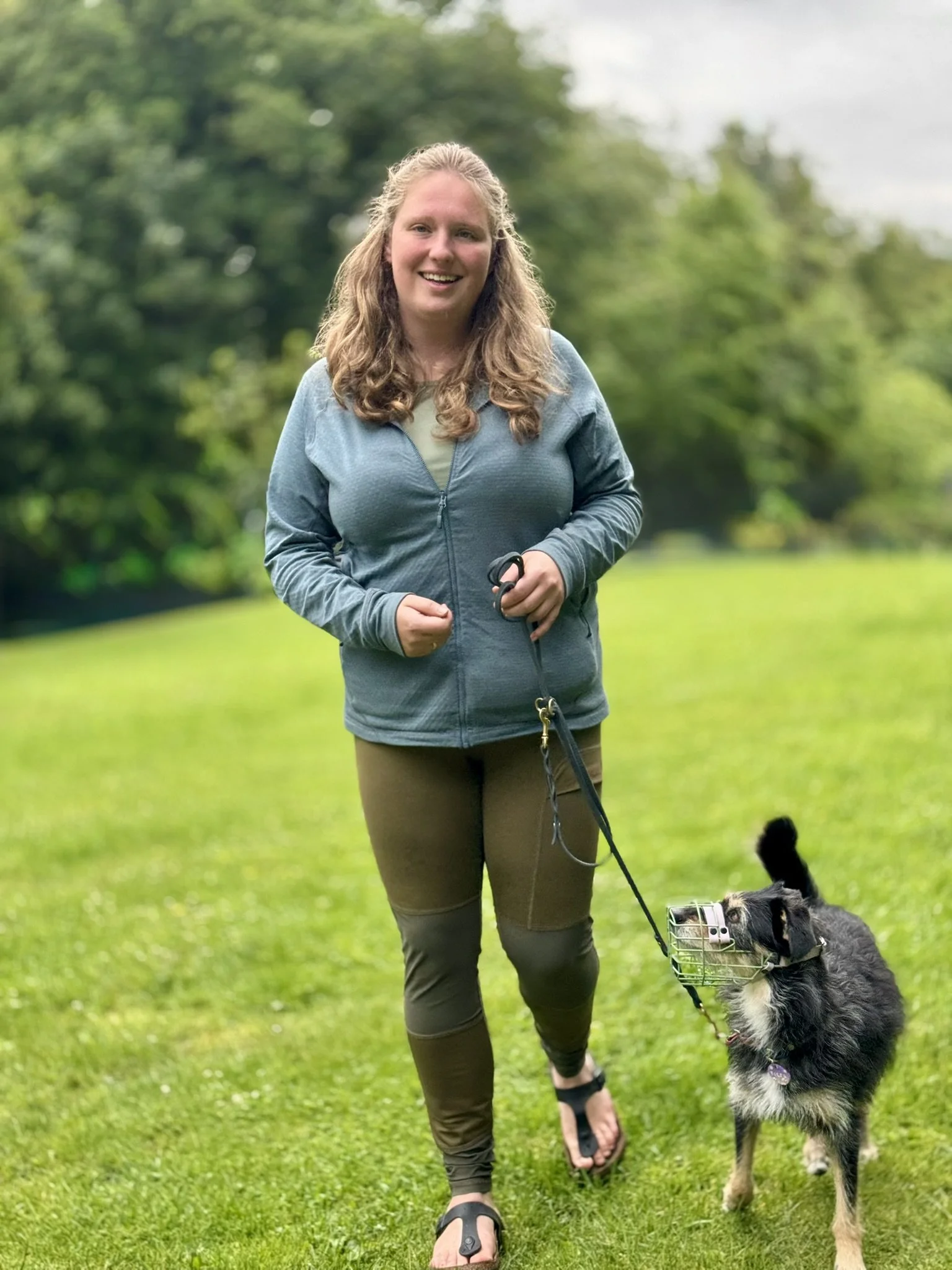 Eine lachende Frau im Freien mit ihrem Hund, der eine Maulkorb trägt, auf grünem Gras mit Bäumen im Hintergrund.