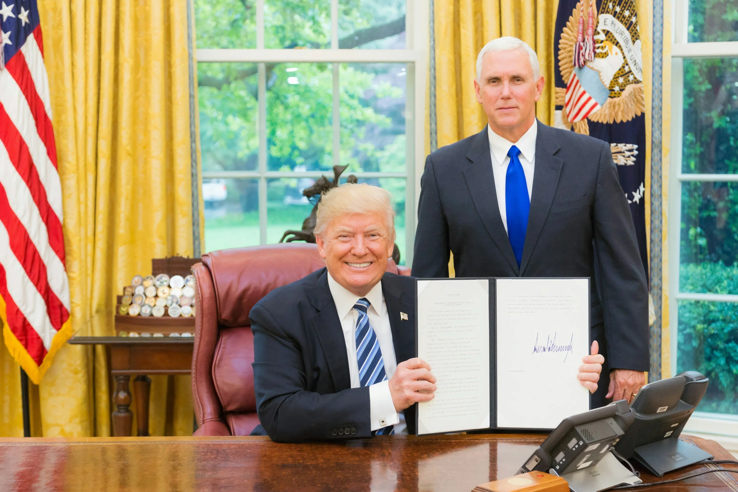 Former President Donald Trump sitting at a desk, holding an open document, in the Oval Office, with a man standing beside him. Trump is smiling and wearing a dark suit, white shirt, and blue striped tie. The man standing is wearing a dark suit, white shirt, and blue tie. The Oval Office has yellow curtains, large windows, and an American flag to the left.