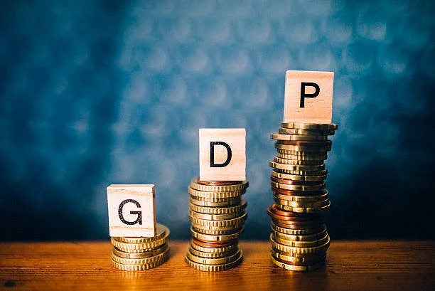Stacks of coins with letter tiles on top spelling 'G', 'D', and 'P' on a wooden surface, representing financial growth or investment levels.