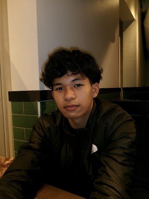 A smiling woman with curly hair, wearing glasses, hoop earrings, and layered necklaces inside a room with red chairs and a window.