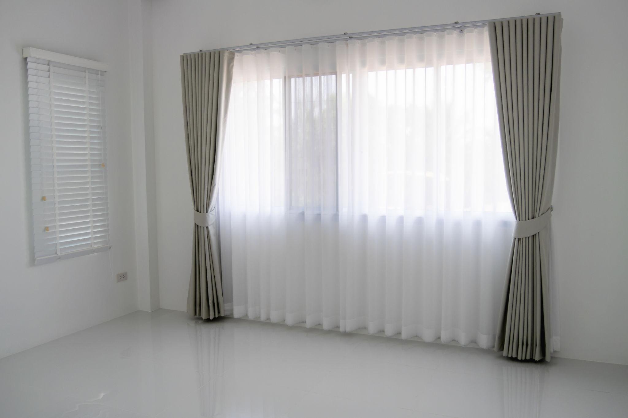 A room with white walls, white floor, a large window with sheer curtains, and a smaller window with white blinds.