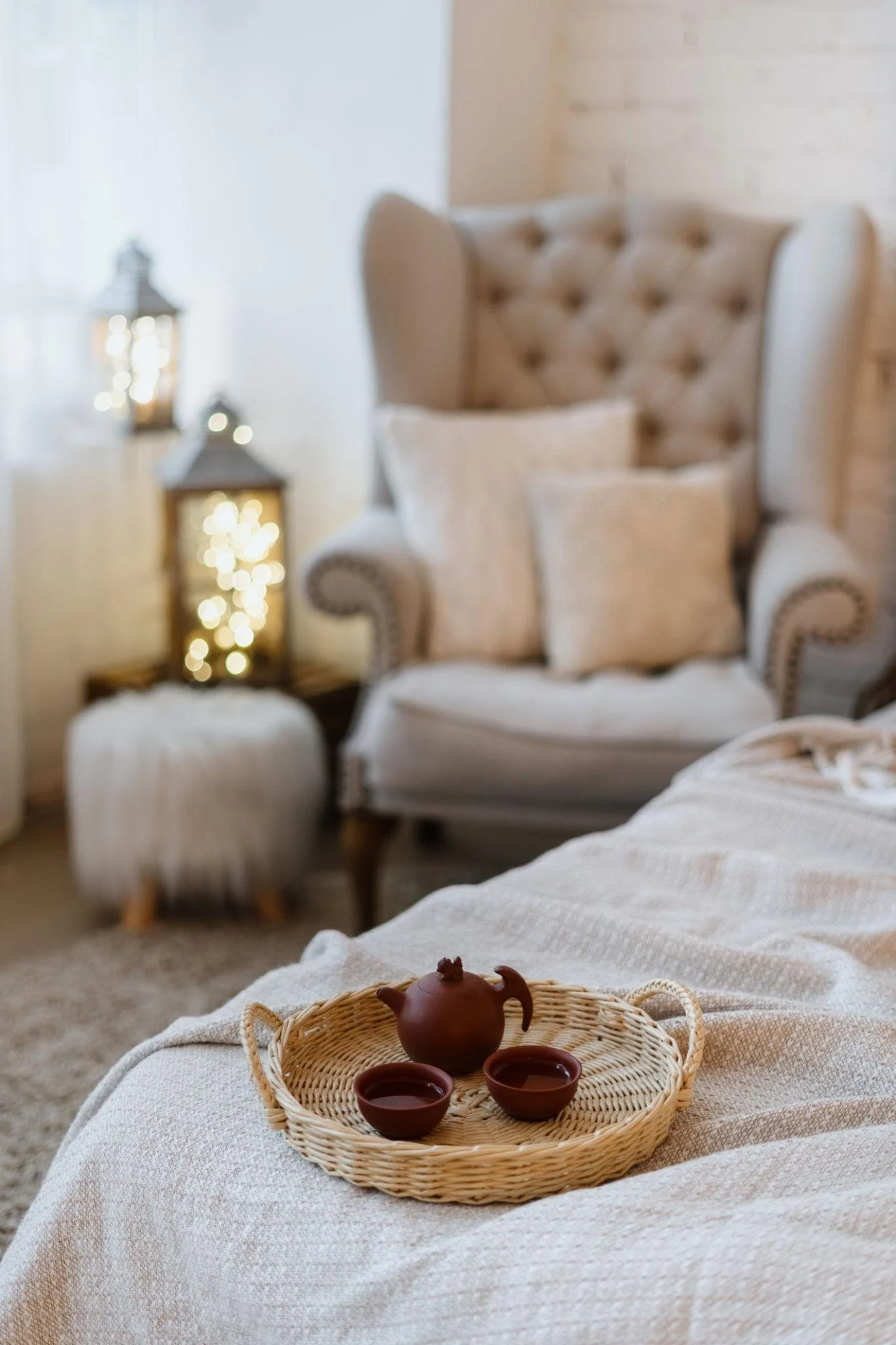 A cozy sitting area with a beige tufted high-back armchair, a matching ottoman, and a small woven tray with a brown teapot and cups on a white textured tablecloth. In the background, two lanterns with string lights are visible, creating a warm and inviting atmosphere.