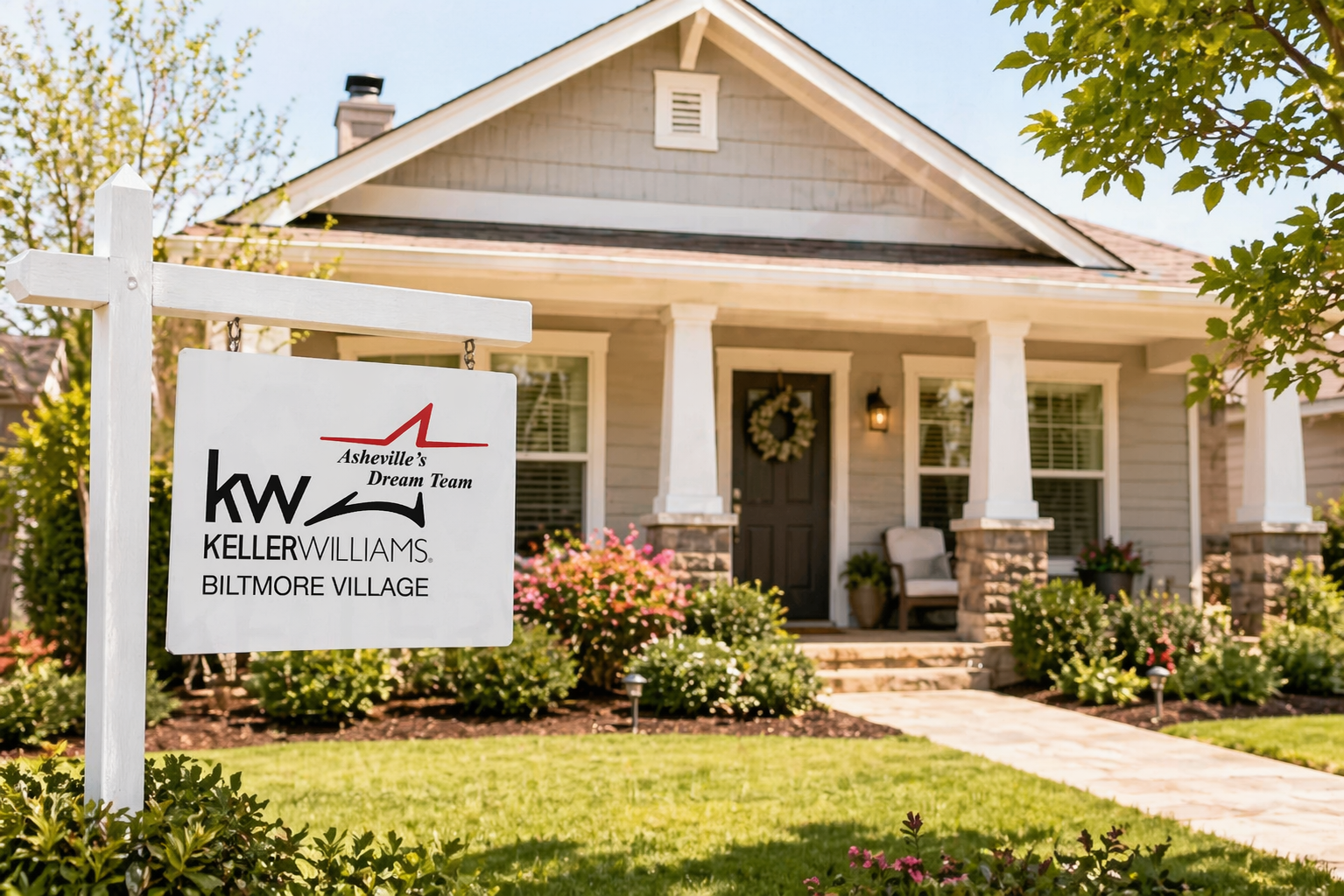 Front yard of a house with a sign for Keller Williams real estate team in Asheville's Biltmore Village. The house has a porch with white columns, a dark front door with a wreath, and flower beds with pink and white flowers.