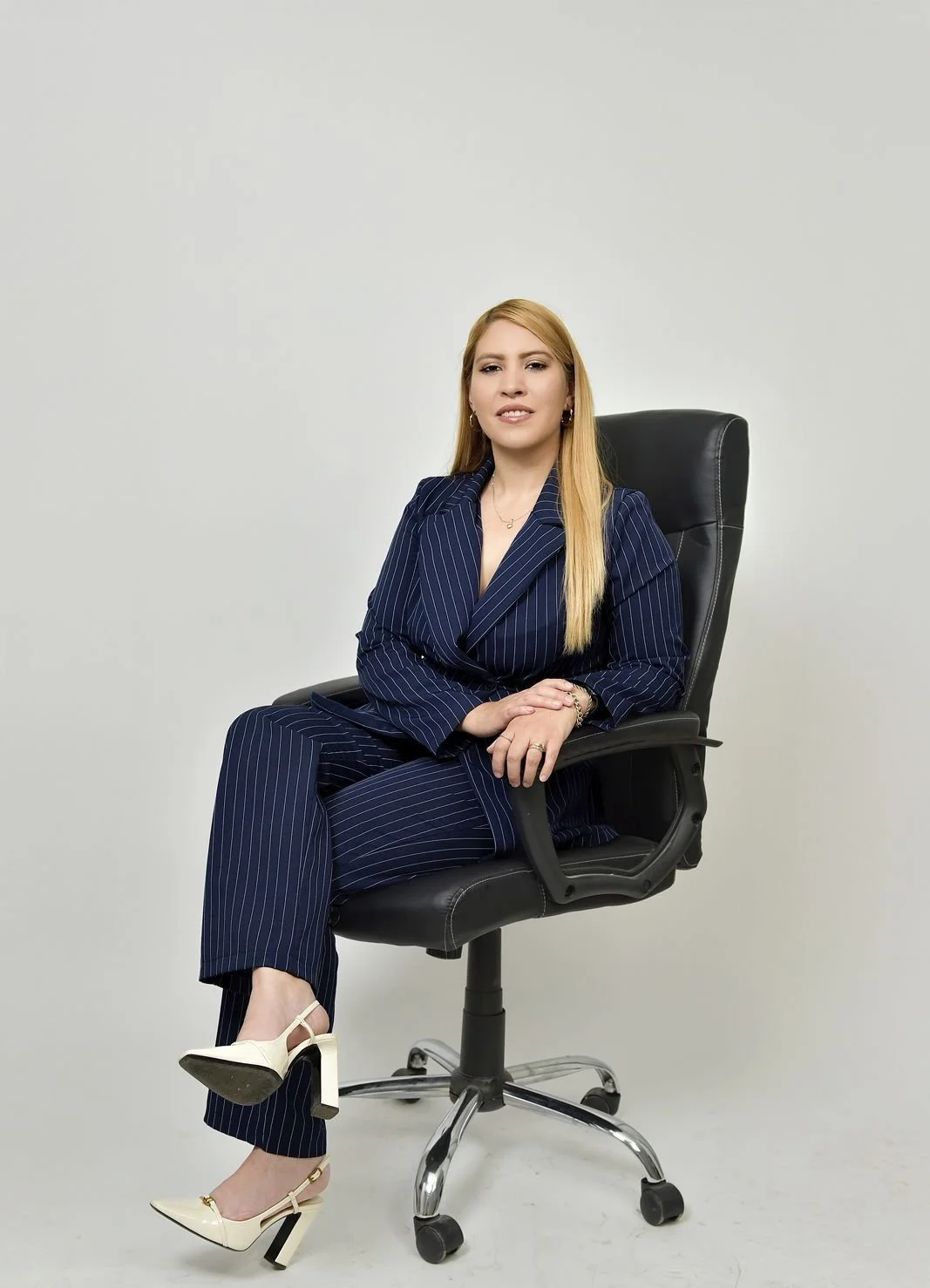Mujer con cabello rubio, usando traje de pinzas azul con rayas, sentado en una silla de oficina negra, con fondo blanco.