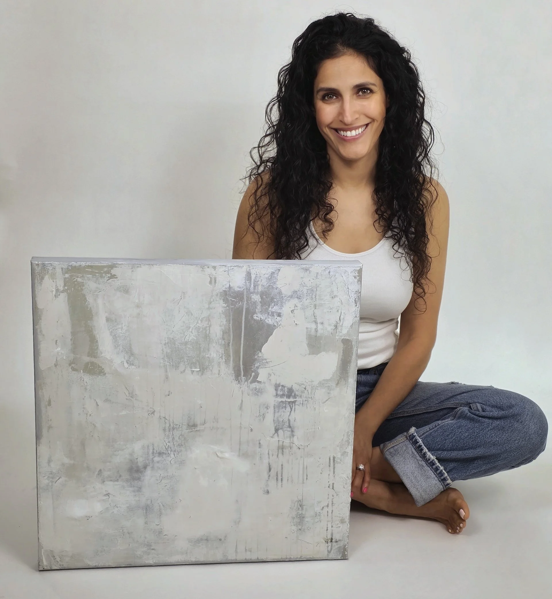 Artist and painter Michelle Lovie seated against a white background holding one of her paintings, a work that combines neutral tones and intricate textures.
