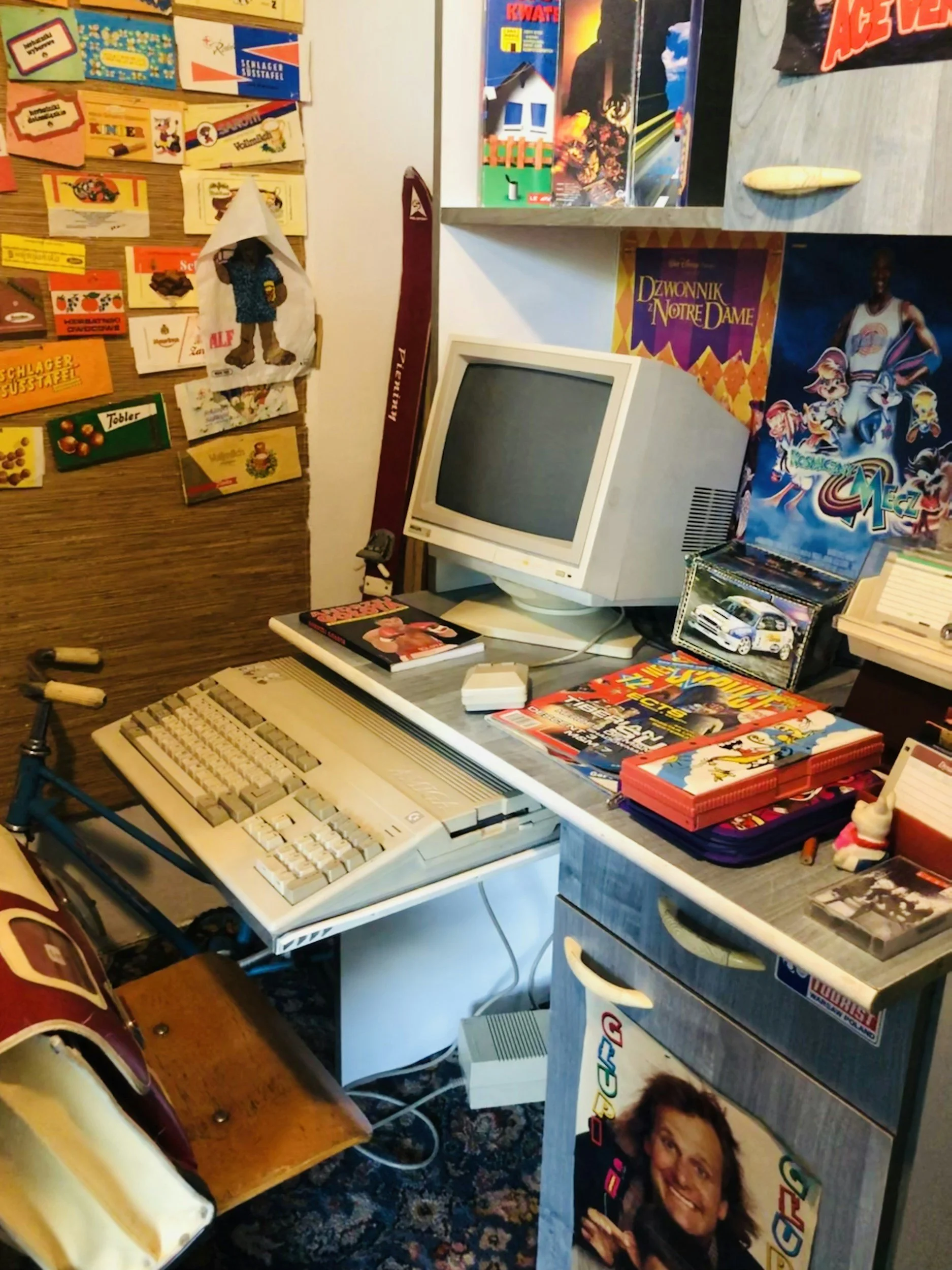 A cluttered vintage computer workspace with a CRT monitor, retro keyboard, and various posters and collectibles on the walls. There are magazines, books, and a small toy on the desk, with a bicycle and patterned rug visible.