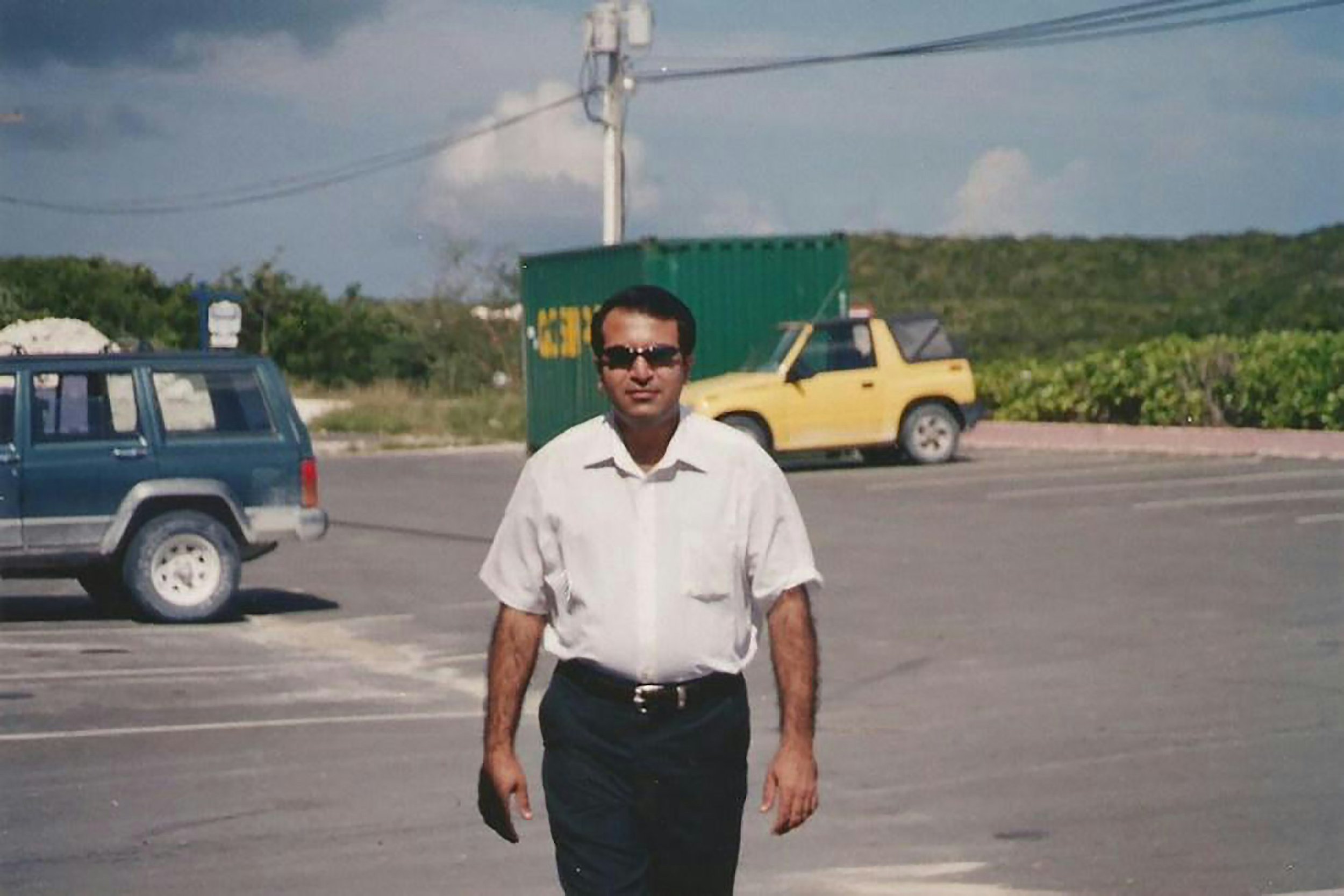 A man wearing a white shirt, black pants, and sunglasses walking in a parking lot with cars and greenery in the background.