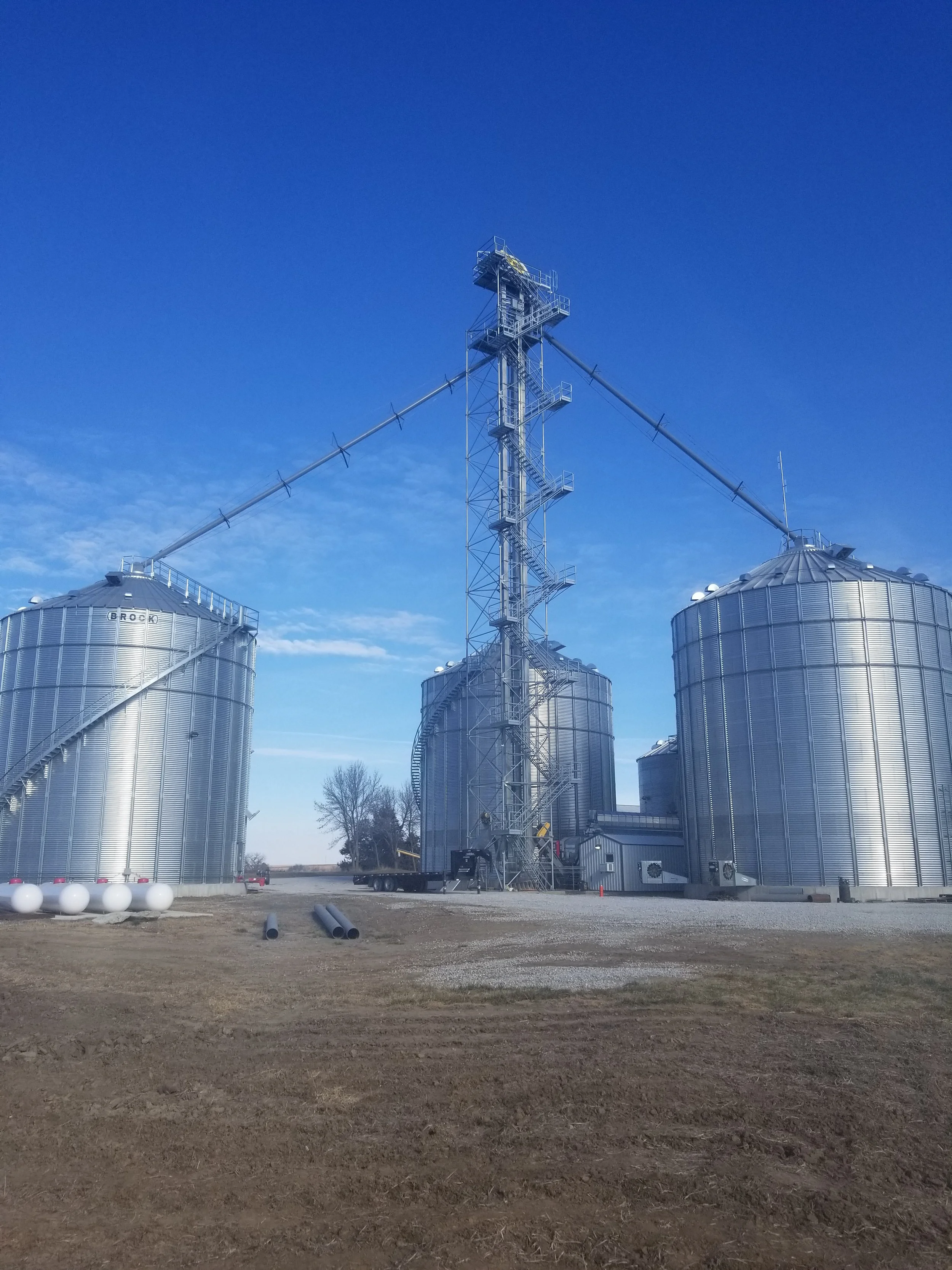 Three large silver grain silos with a tall metal tower in the center, connected by long metal beams, under a clear blue sky.