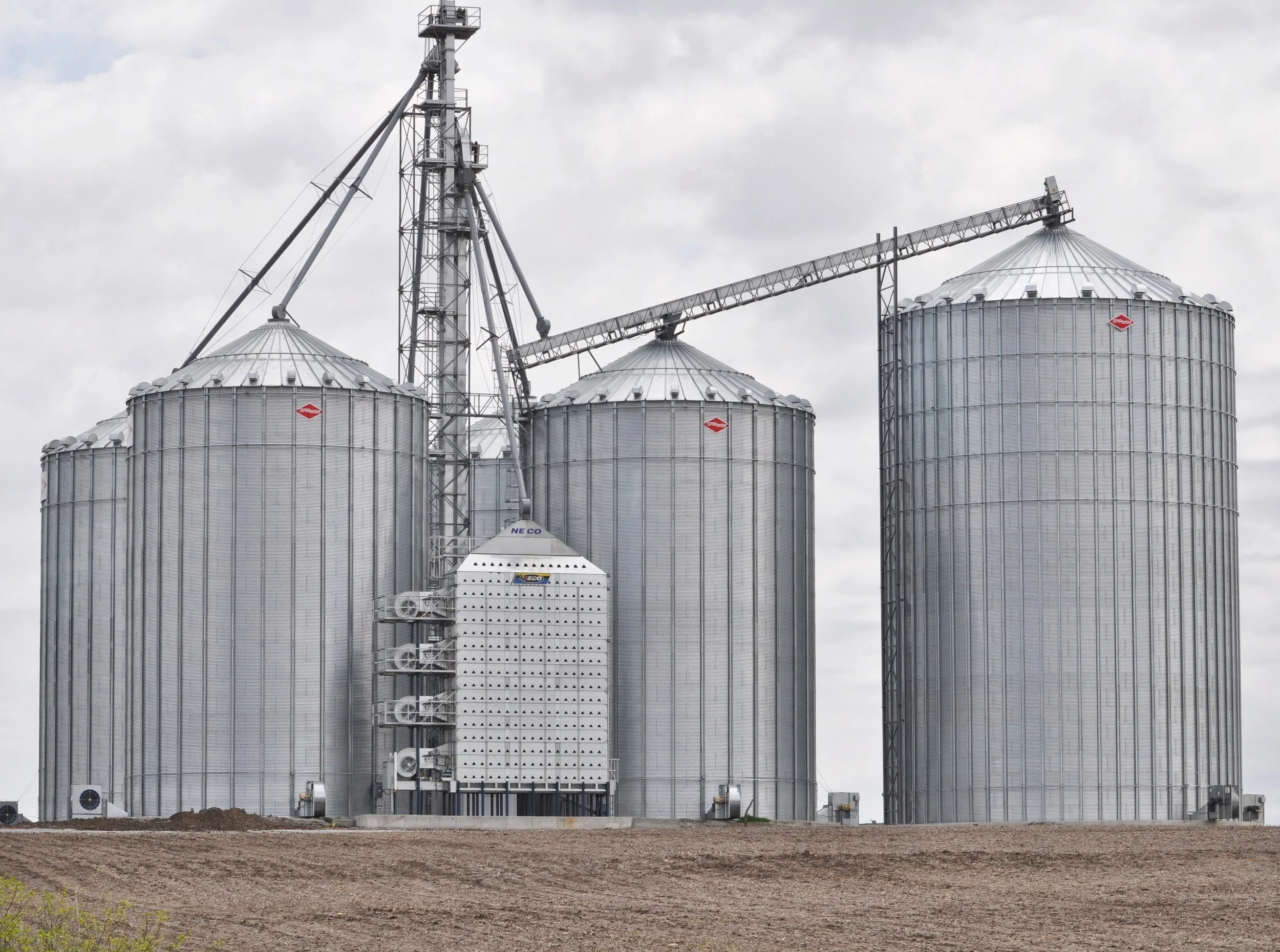 Four large metal grain silos with a crane and ladder structure on a cloudy day on farmland.