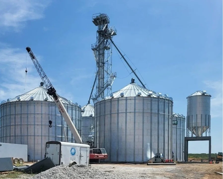 Industrial grain silos with a crane and construction equipment, under a blue sky.