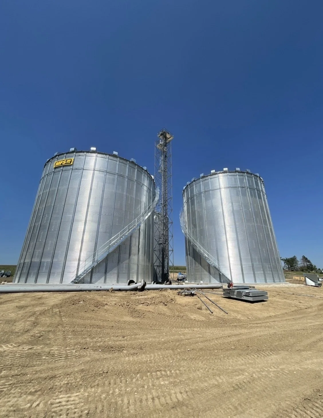 Two large metal grain silos with spiral staircases, situated on a dirt site under a clear blue sky.