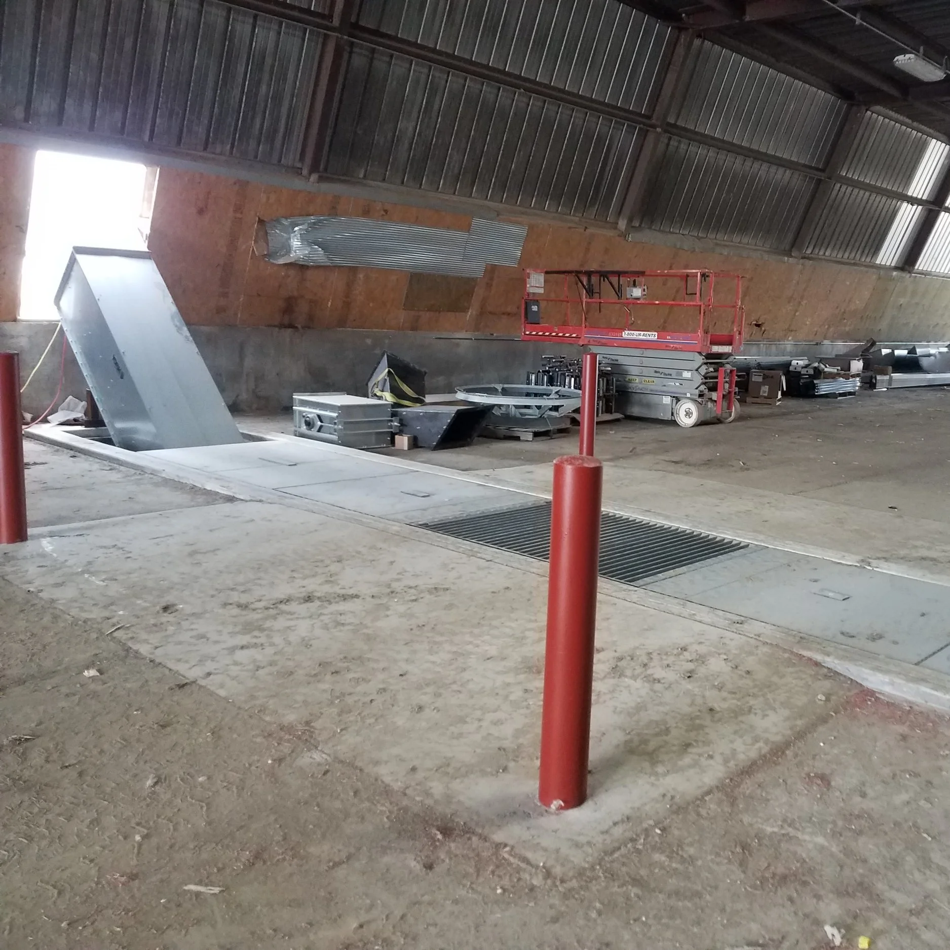 Construction site with a partially installed escalator, scissor lift, and construction tools inside a large building with sloped wood-paneled walls and a concrete floor.