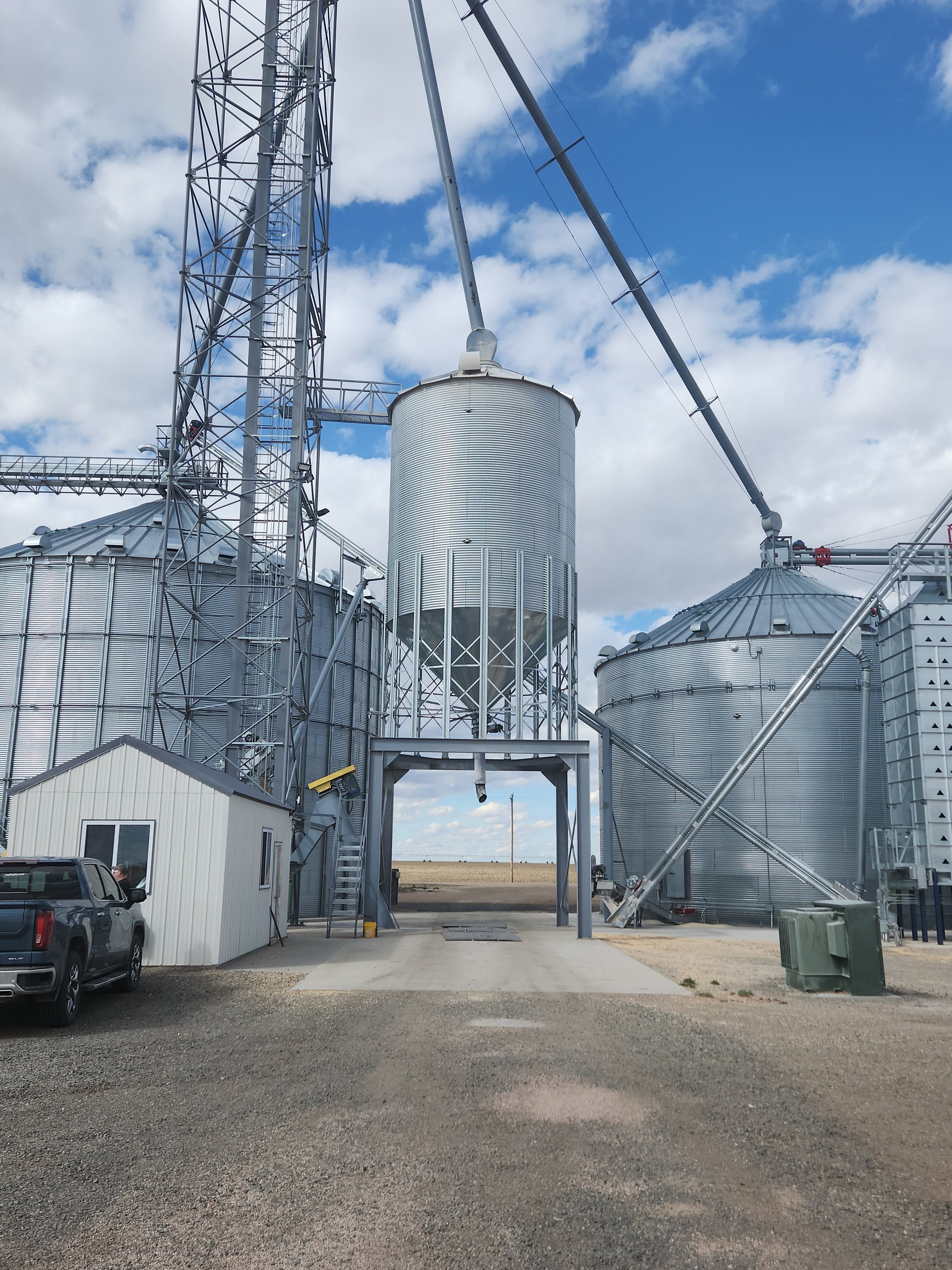 A grain storage facility with metal silos and a tall grain elevator structure under a partly cloudy sky.