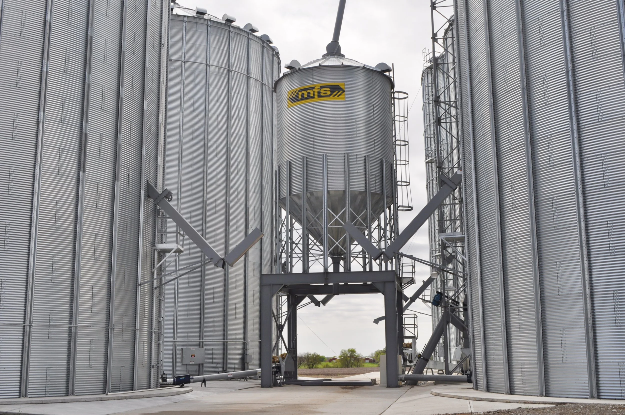 Large industrial grain storage silos with metal exteriors, connected by walkways and ladders, under a cloudy sky.