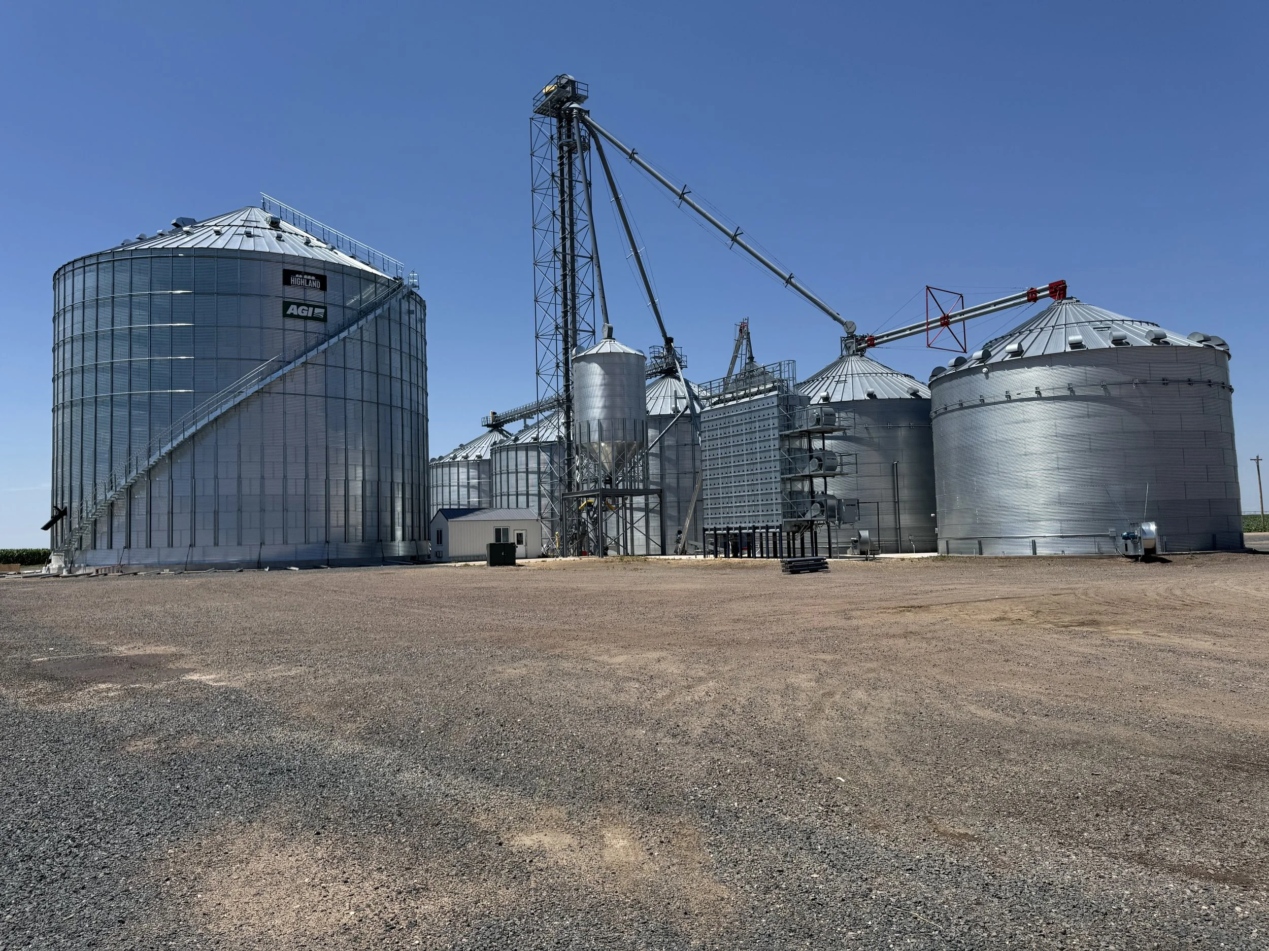 A row of large silver grain silos and storage tanks on a farm under a clear blue sky, with gravel ground in the foreground.