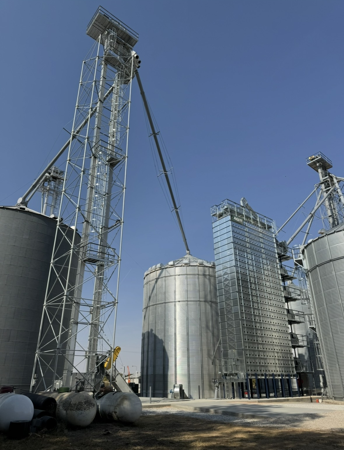 Industrial grain silos and storage tanks with tall metal structures and ladders against a blue sky.