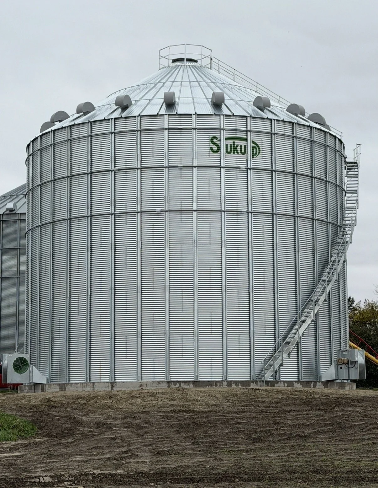 Large gray metal grain silo with the word 'SUKUP' on it, exterior staircase on the right, and a cloudy sky above.