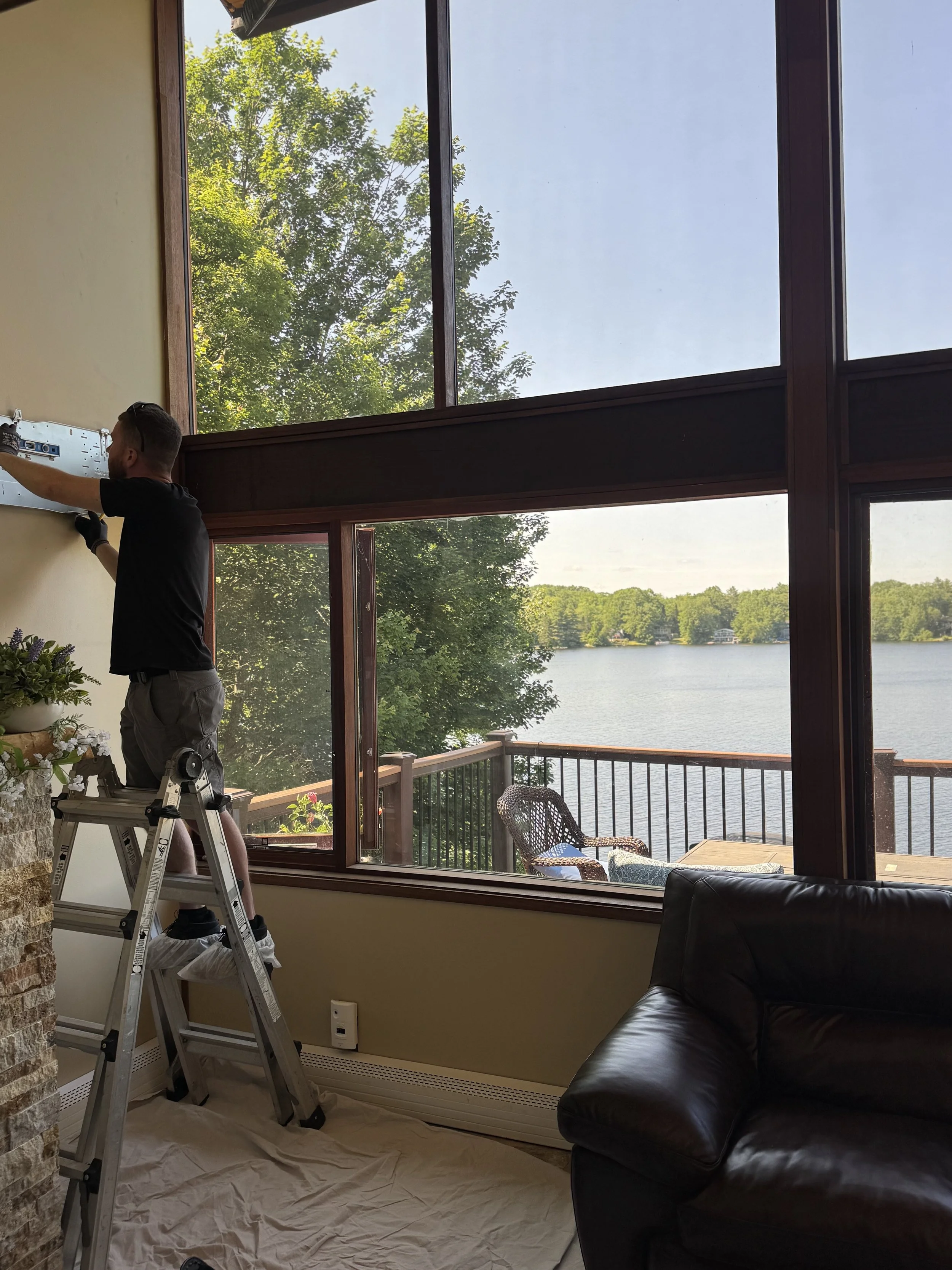 A man standing on a ladder installing or working on a rack or shelf on a wall inside a room with large windows overlooking a lake and trees outside.