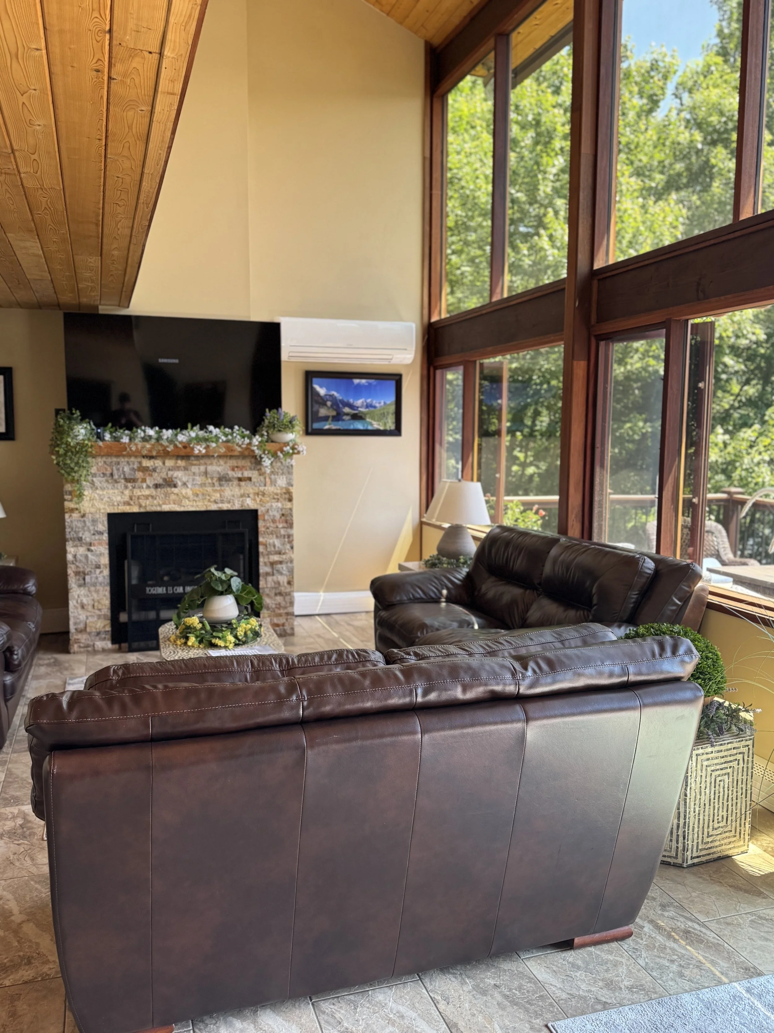 Living room with large windows, leather sofas, a fireplace with a stone surround, and a flat screen TV above it, with sunlight streaming through the windows.