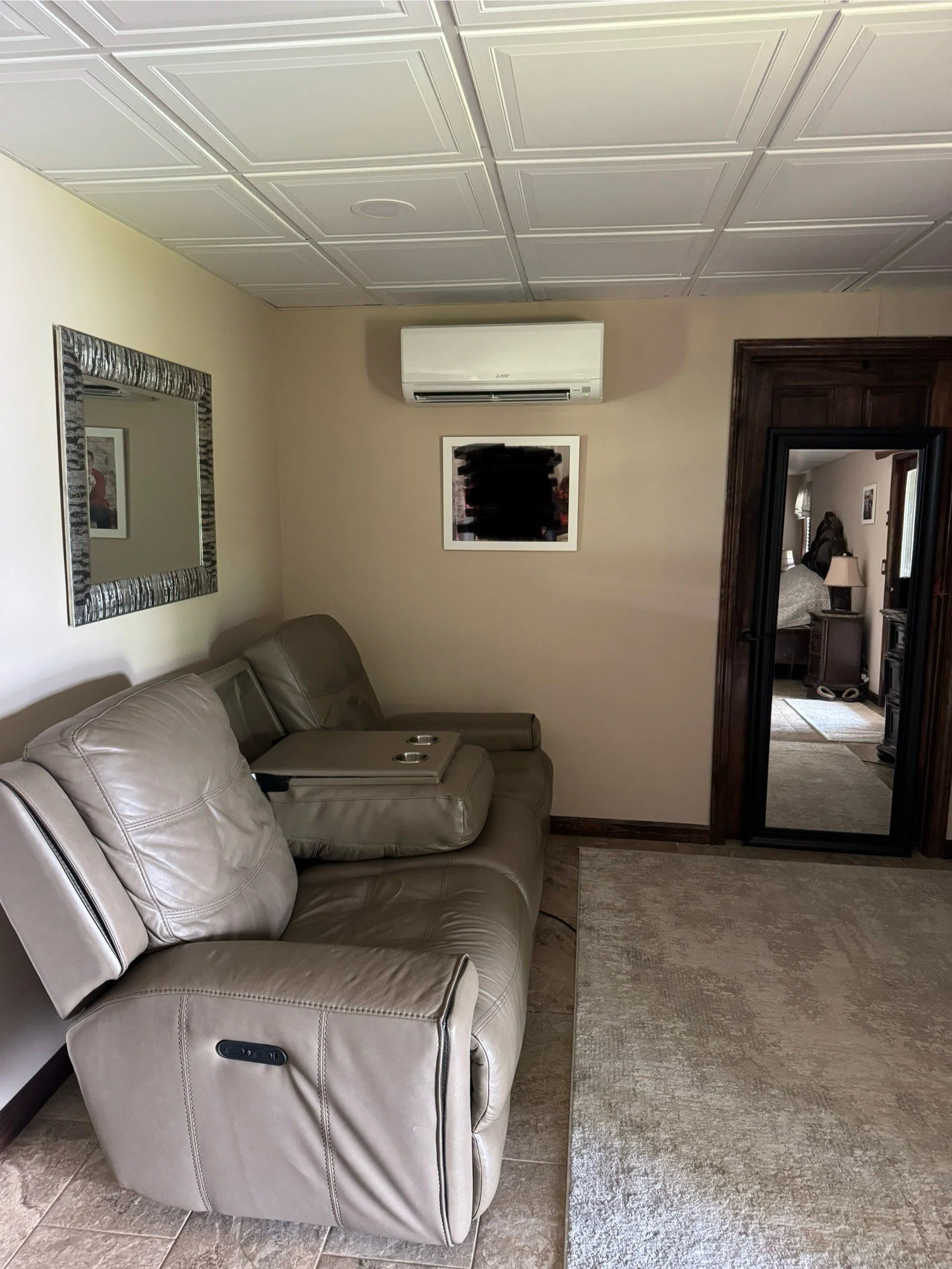 A living room corner with a beige leather sofa, a wall-mounted air conditioning unit, a framed picture with blacked-out content, a large mirror, and a doorway leading to a bedroom.