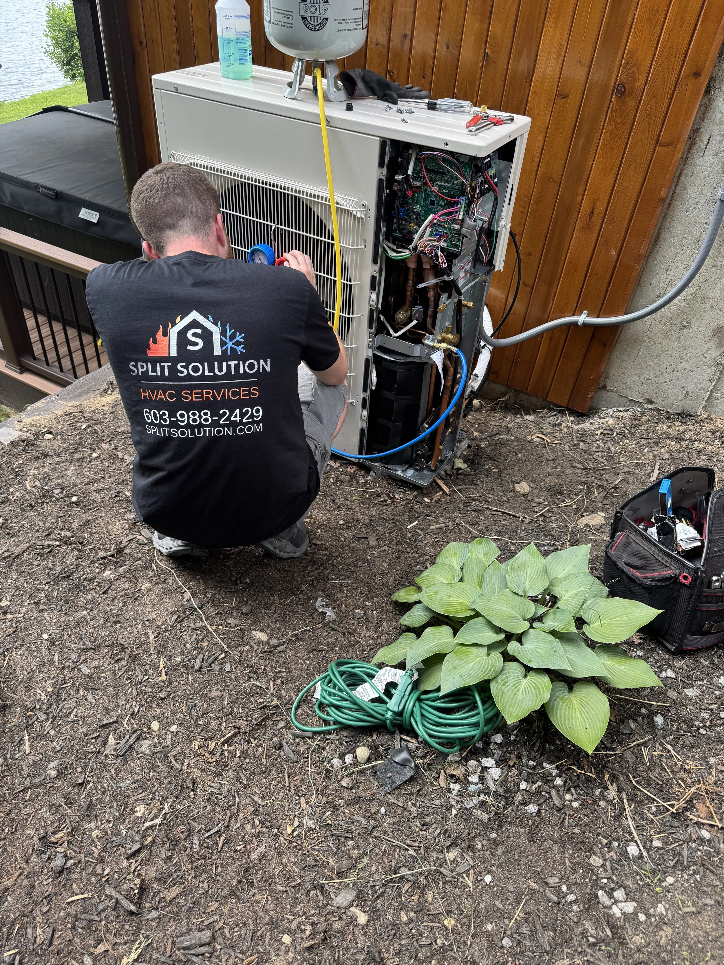 A technician working on a split air conditioning unit outside, with tools, a black toolbox, a green extension cord, and a potted plant nearby.