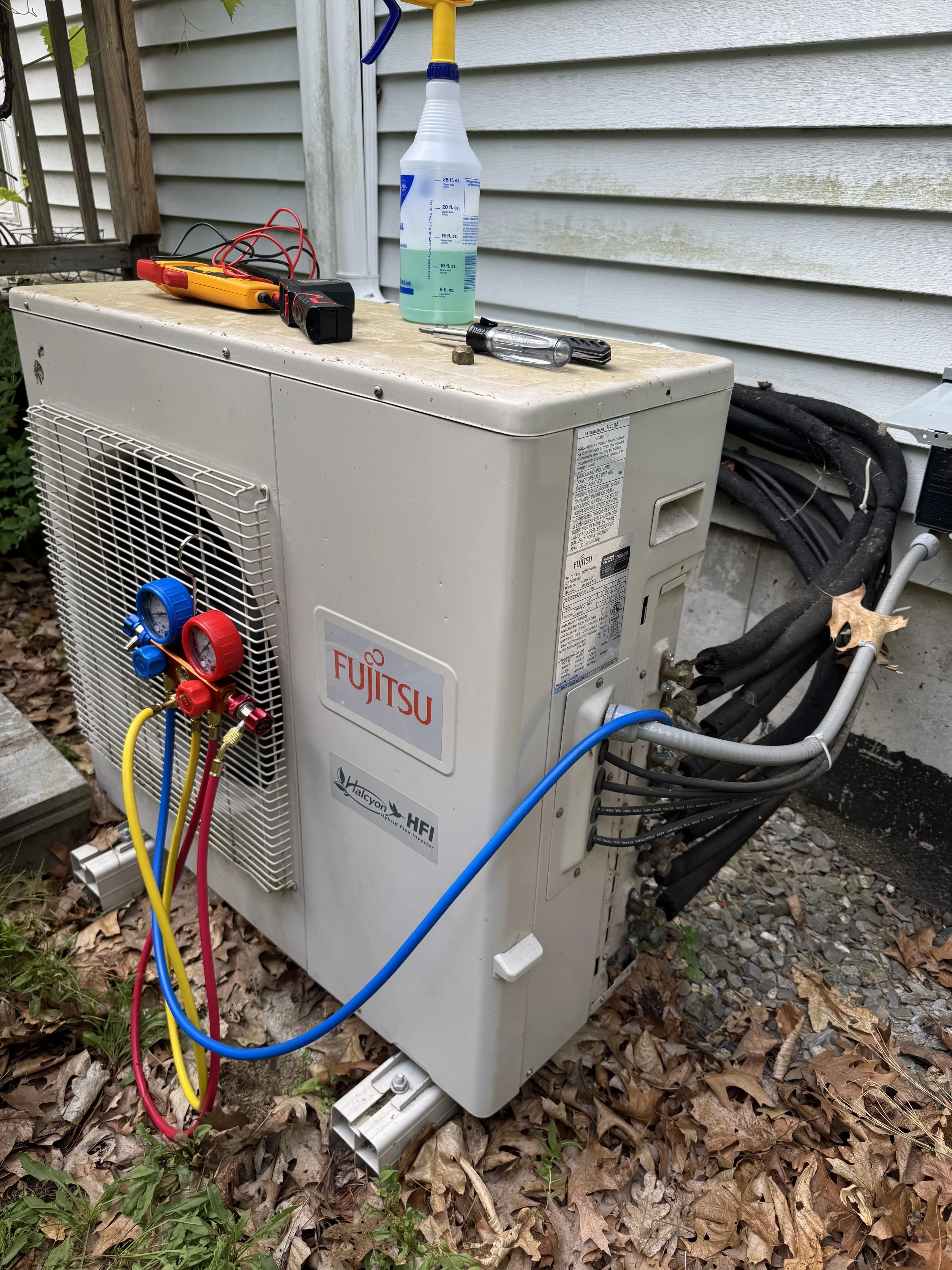 Outdoor Fujitsu HVAC unit with gauges, multimeter, spray bottle, and screwdriver on top. Worn leaves on the ground and electrical wiring connected to the unit.