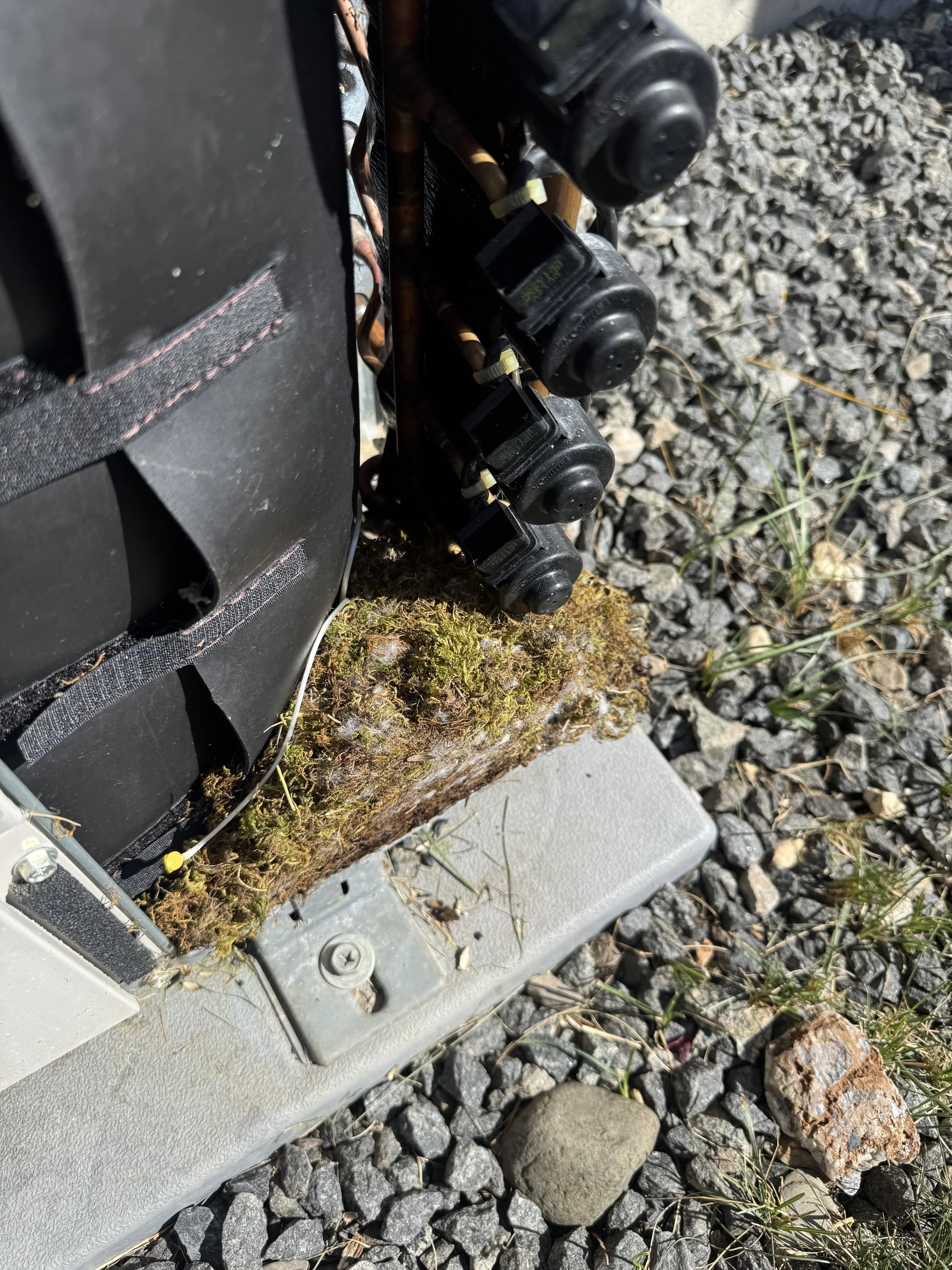 Close-up of a piece of outdoor electrical or telecommunications equipment mounted on a metal base, with black components and copper wiring, on a gravel surface with grass and rocks nearby.
