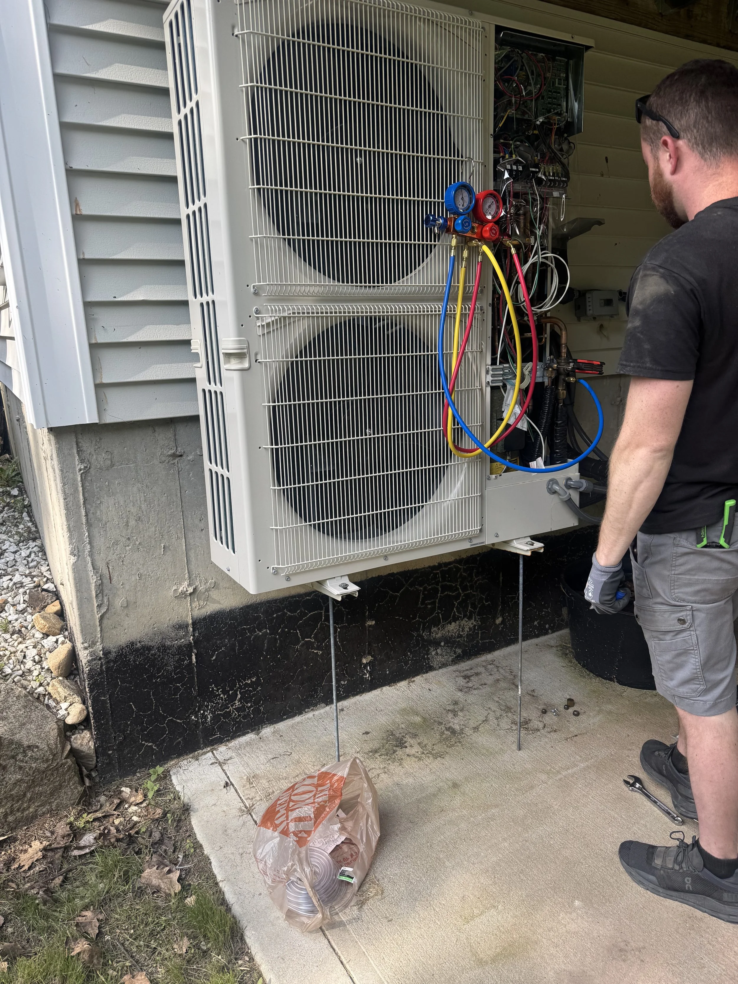 Technician working on a large outdoor air conditioning unit, which has its cover removed to reveal the internal components and refrigerant hoses.