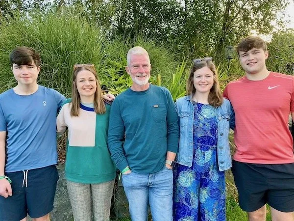 A group of five people standing outdoors in front of green bushes, smiling at the camera. They appear to be a family or friends, with two young men, two young women, and an older man, all dressed casually.